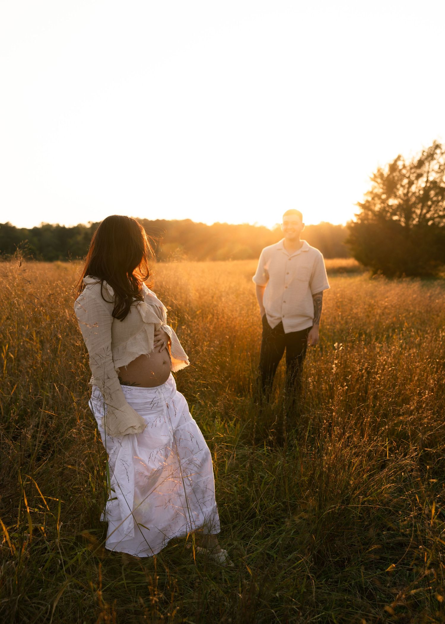 An expectant mama and her husband gaze loving at each other in a wheat field at sunset