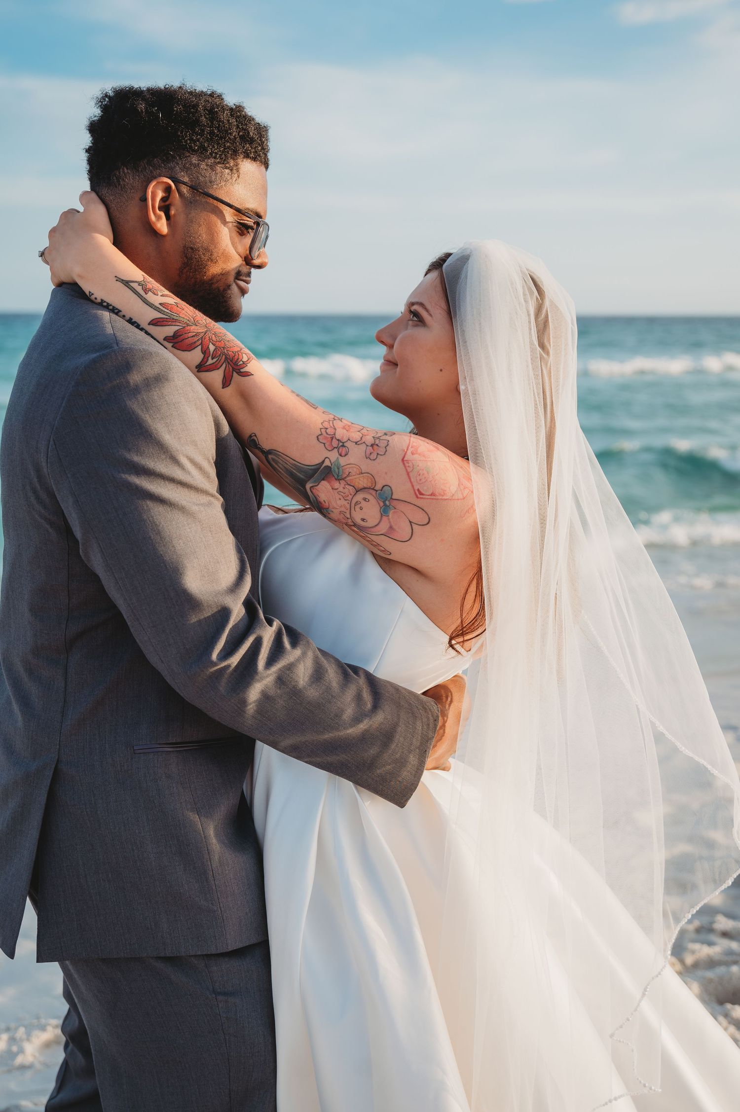 Bride and groom embrace on 30A beach, she wears white gown and veil with floral tattoos, he wears gray suit for wedding.