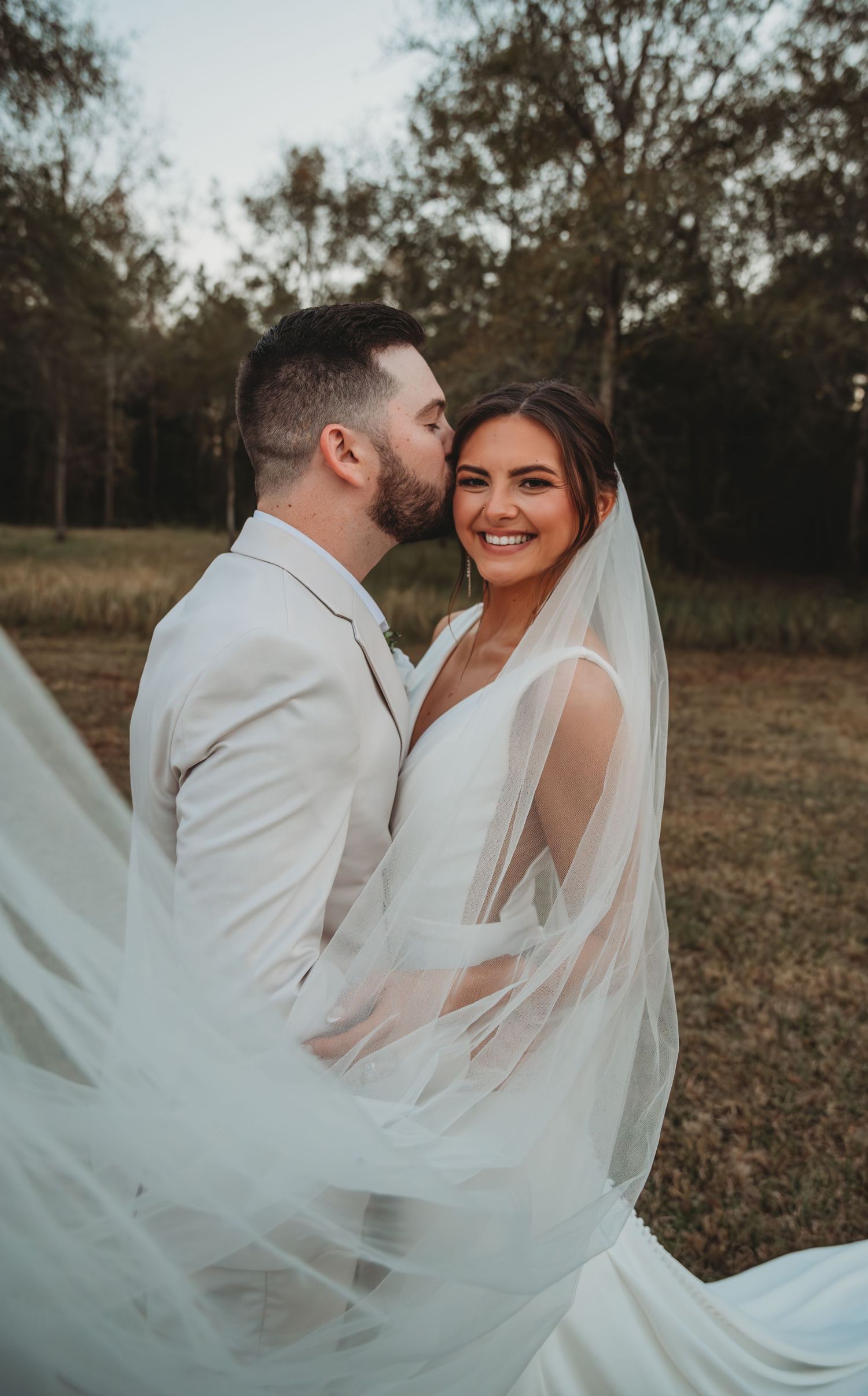 A romantic outdoor Panama City Beach wedding photo with flowing white veil and light colored suit against a natural wooded backdrop.