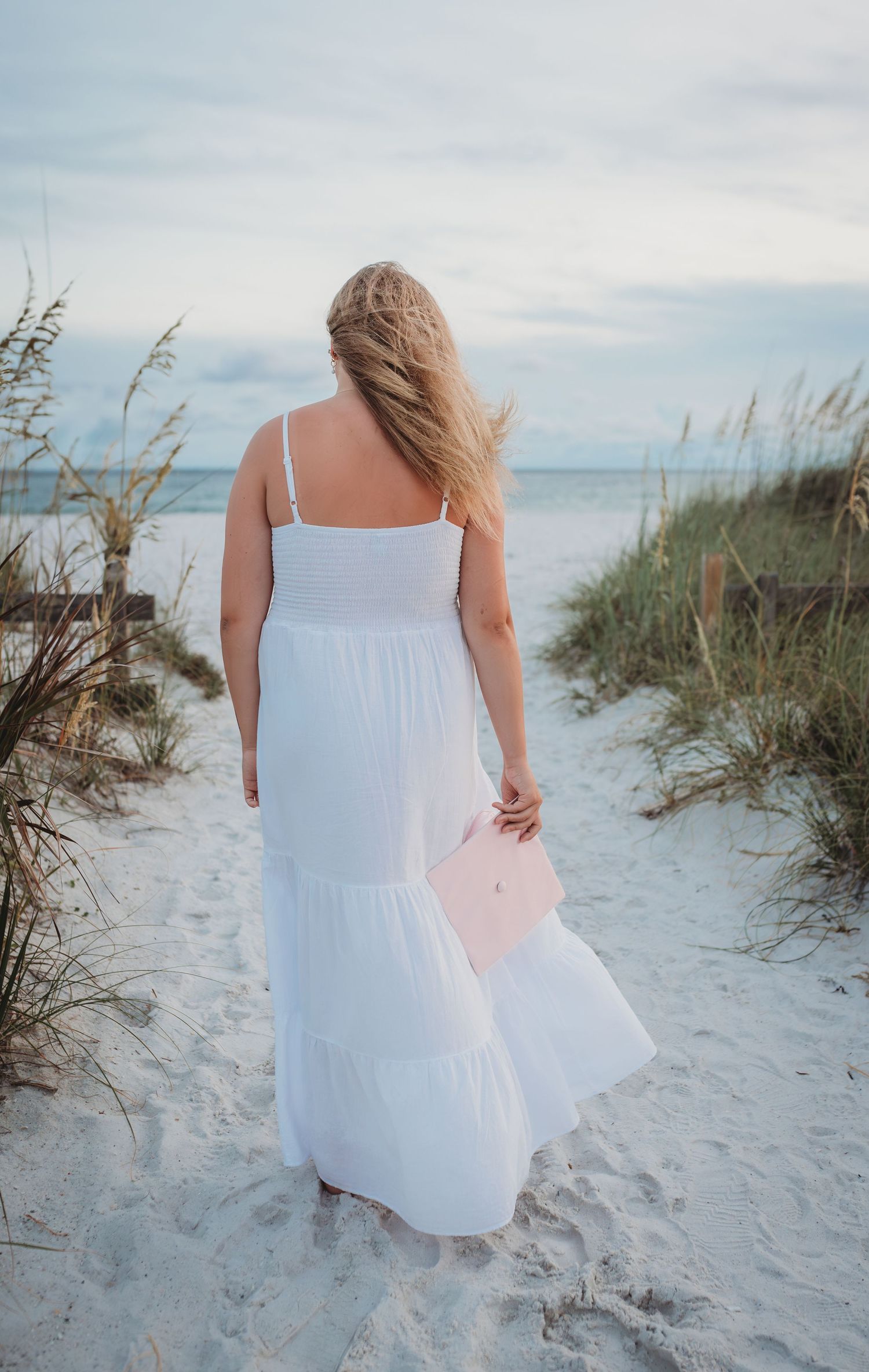 A senior in a flowing white sundress walks along a sandy beach path through sea oats at sunset holding a graduation cap in Panama City Beach