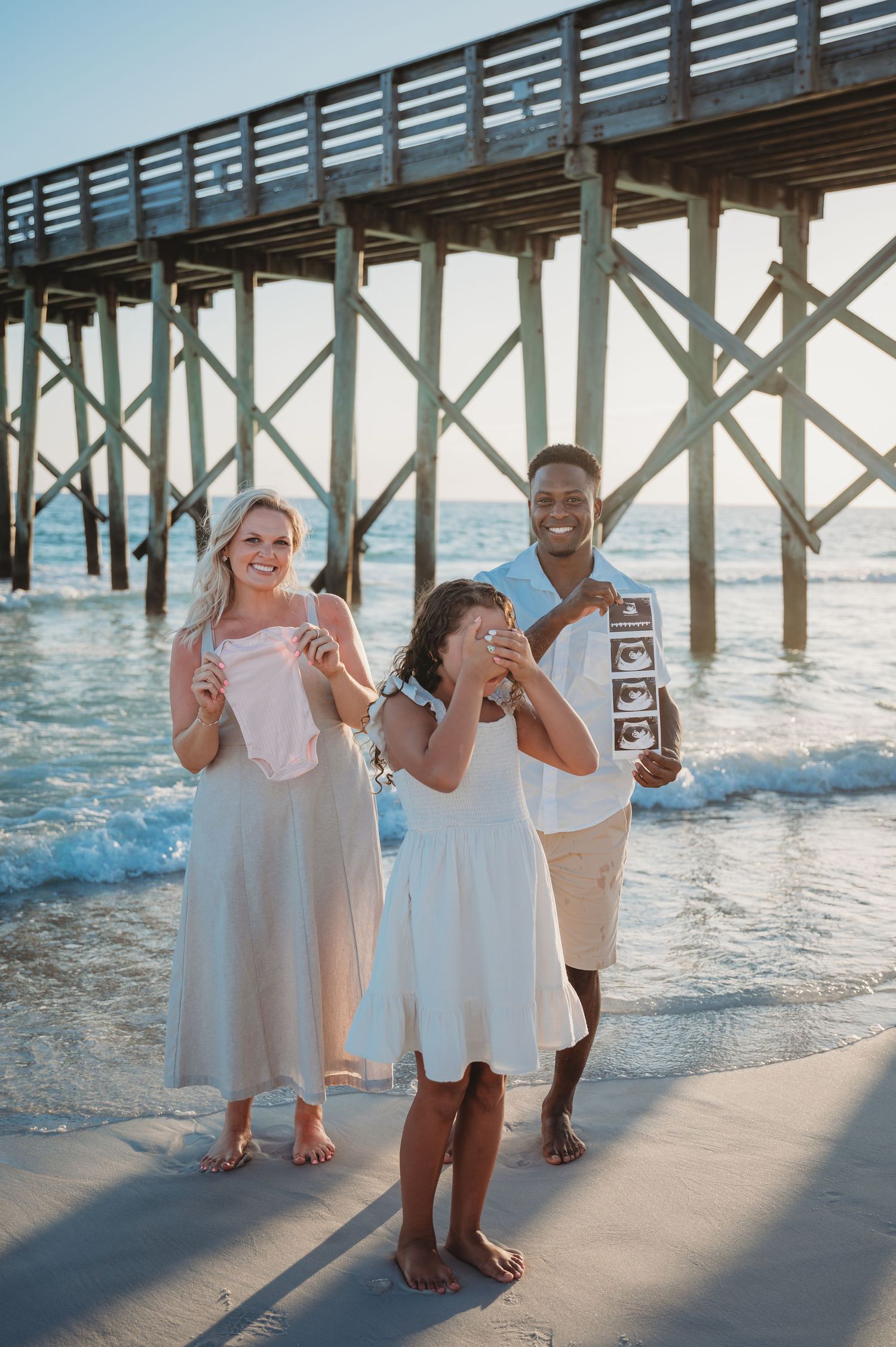A joyful beach moment captured in front of a wooden pier with people in white outfits sharing a pregnancy announcement in Panama City Beach.