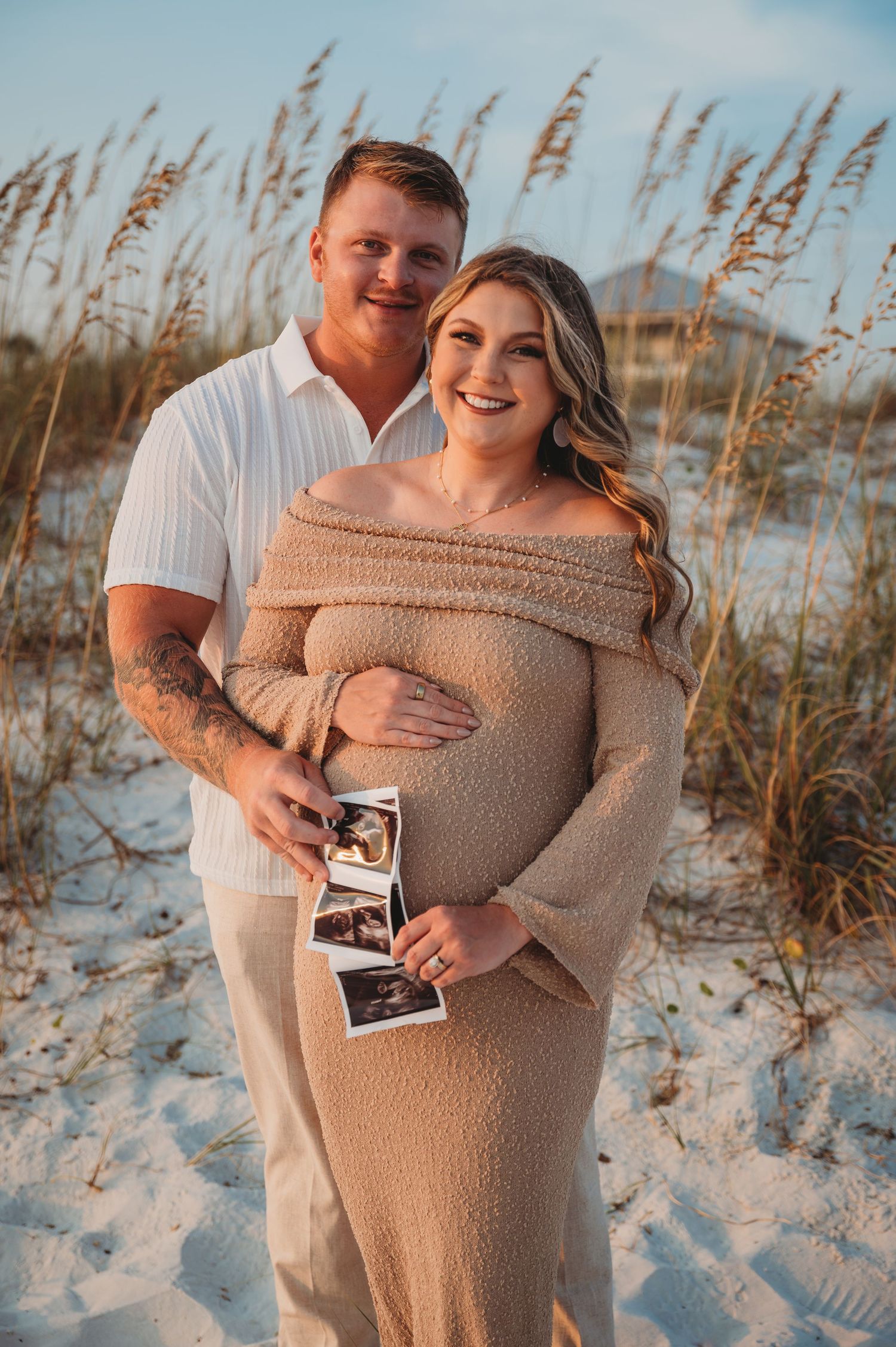 A couple poses on a beach at sunset holding ultrasound photos, with sea oats swaying in the golden light in Panama City Beach.