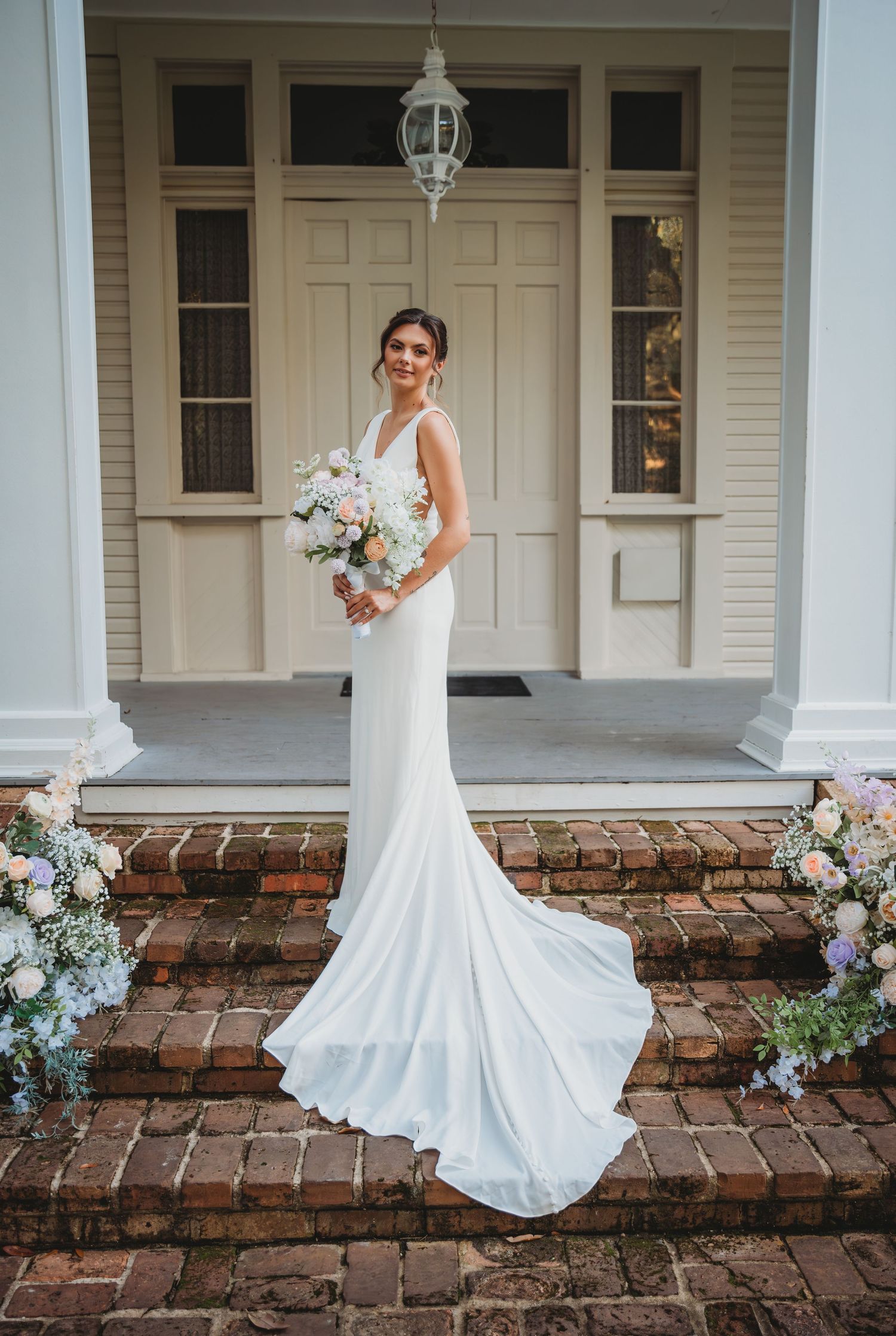 A bride in a wedding gown poses on a brick porch with white columns and floral arrangements during a Panama City Beach wedding.