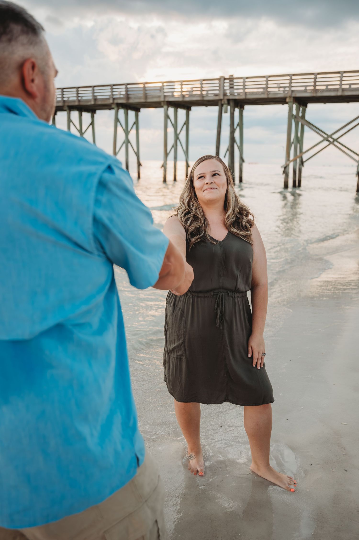 Person in dark dress poses playfully on sandy beach near wooden pier during cloudy day at the ocean in Panama City Beach.