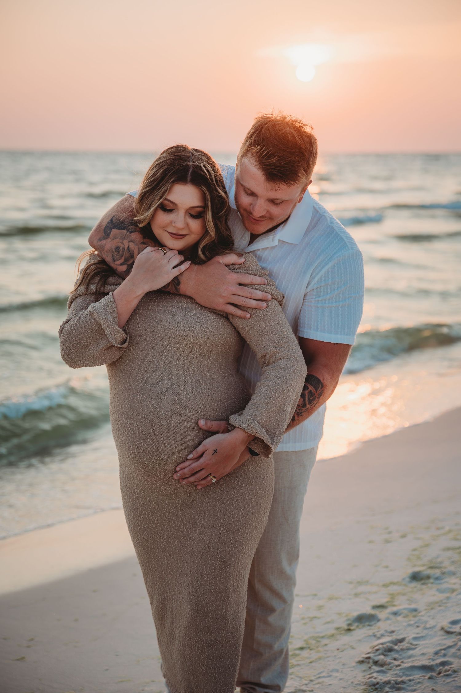 A couple embraces on a beach at sunset, with the pregnant figure silhouetted against the golden sky and ocean waves in Laguna Beach.
