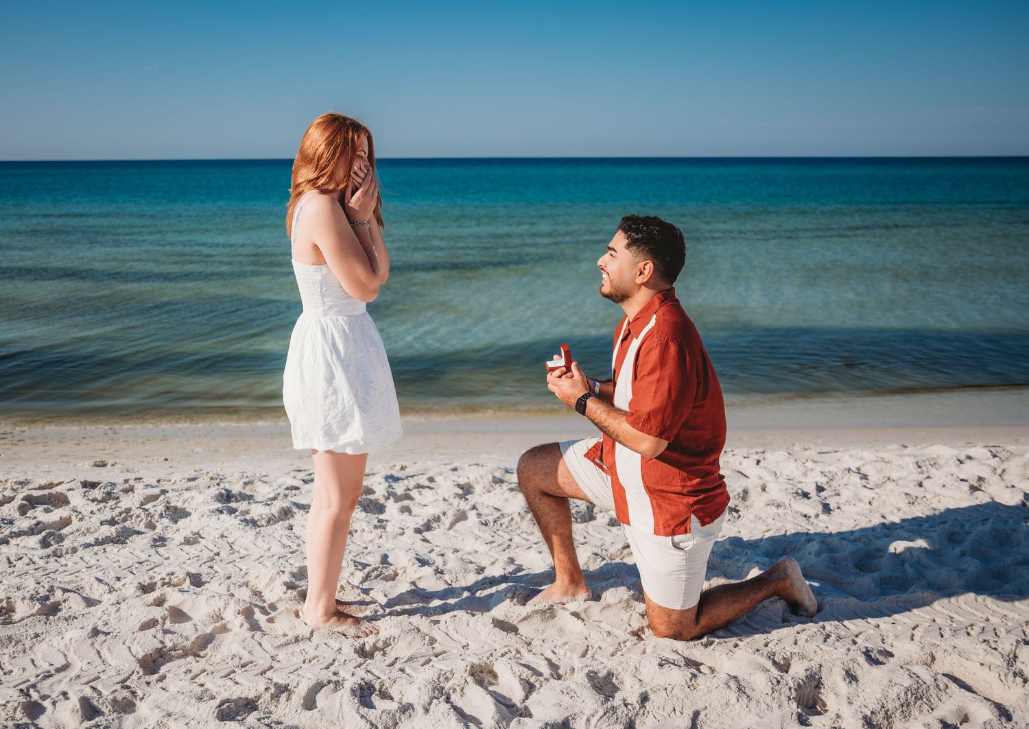A romantic surprise beach proposal scene with a man kneeling before a girl in white by turquoise ocean waters in Panama City Beach.