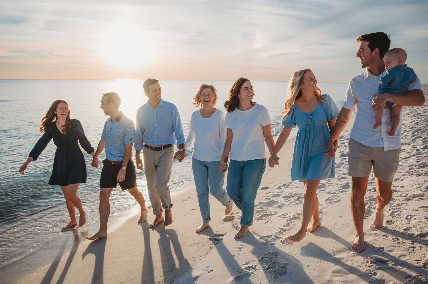 A group of people walking together along a Emerald Coast beach at sunset, holding hands and casting long shadows on the sand.