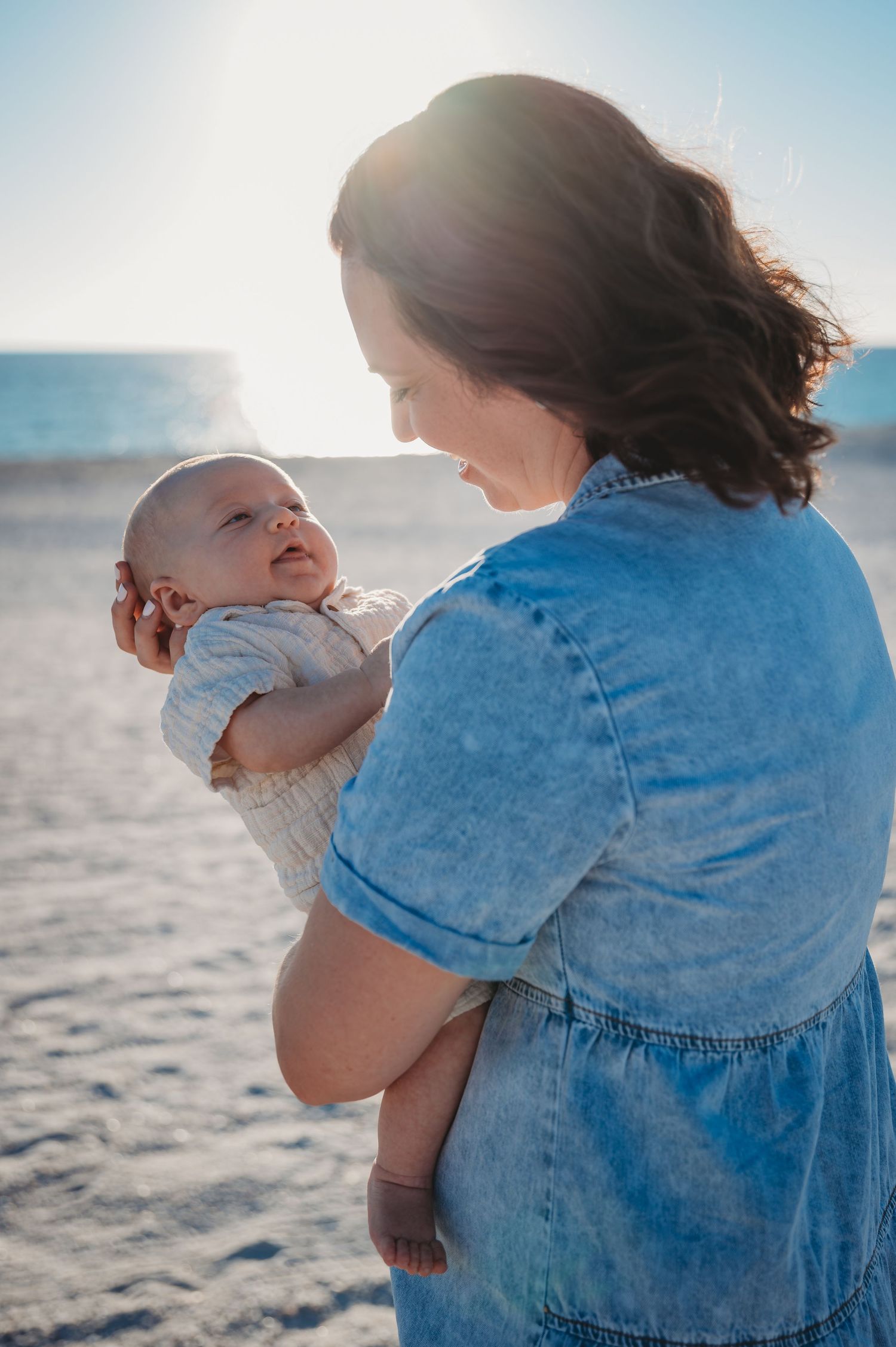 Parent in denim shirt holds baby against bright sunlit St. Andrew's beach backdrop creating tender silhouette moments.