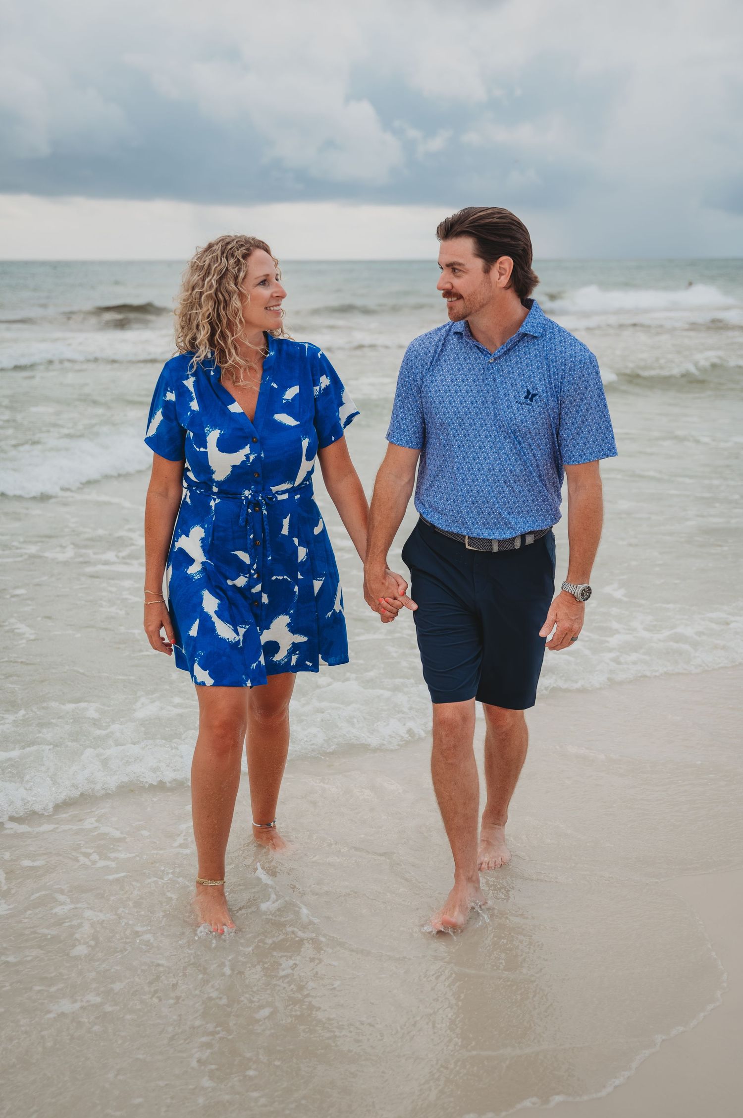 A couple walking hand in hand along a beach shoreline in coordinating blue outfits as waves roll in in Panama City Beach.