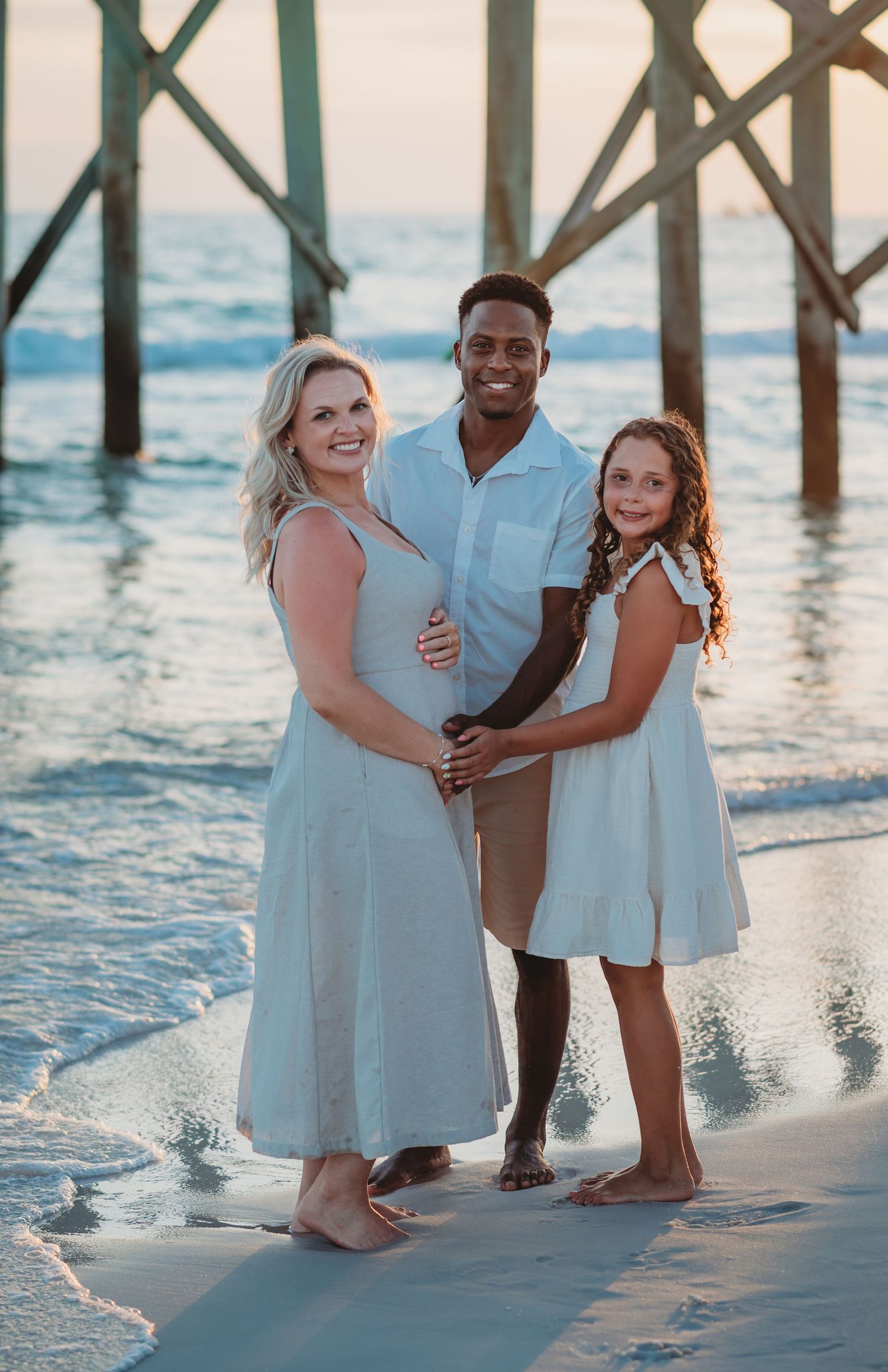A family wearing coordinating white outfits poses together at sunset on a beach beneath a wooden pier in Panama City Beach.