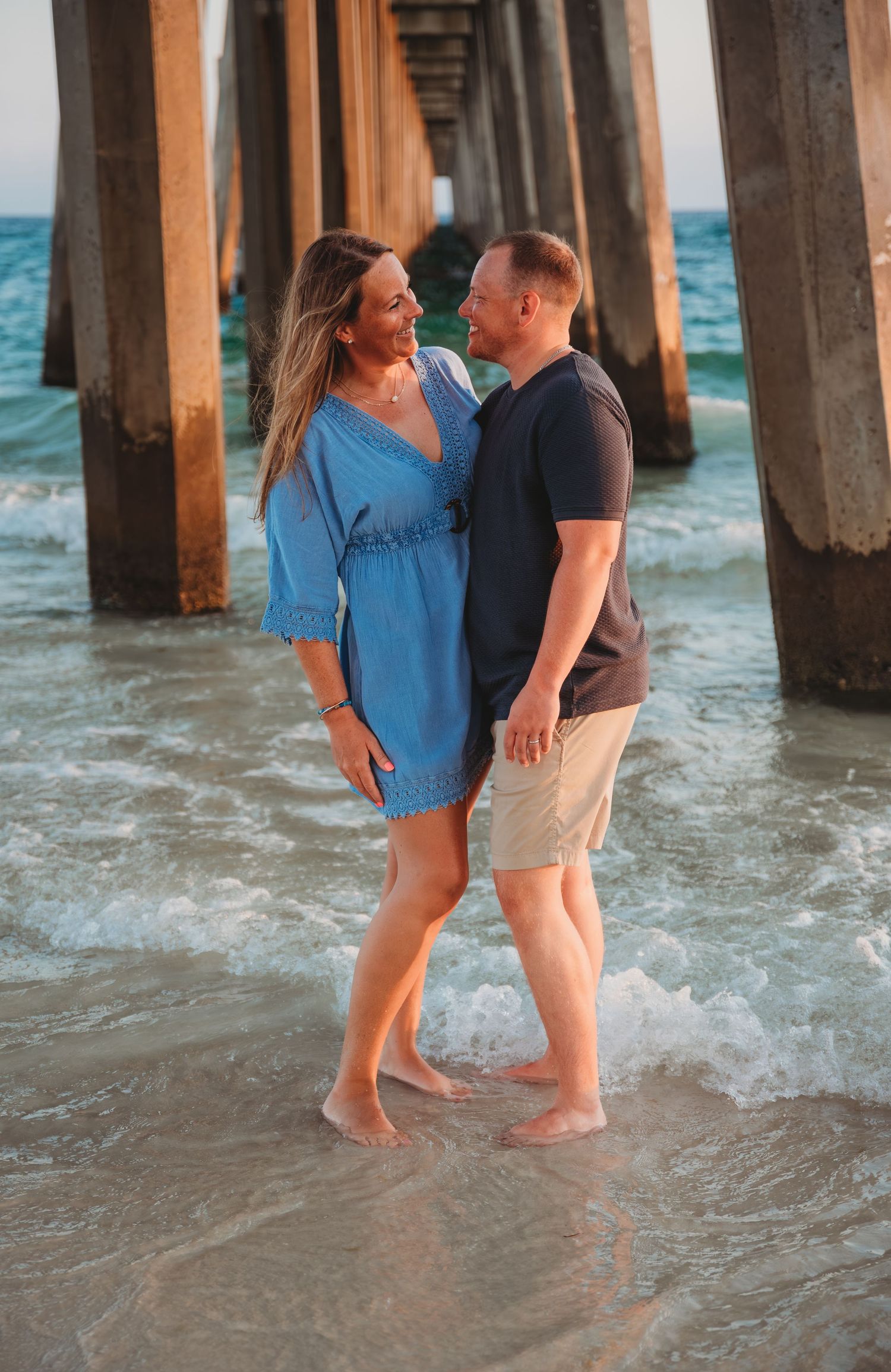 A couple shares a romantic moment standing in shallow ocean water beneath a wooden pier at sunset in Panama City Beach.