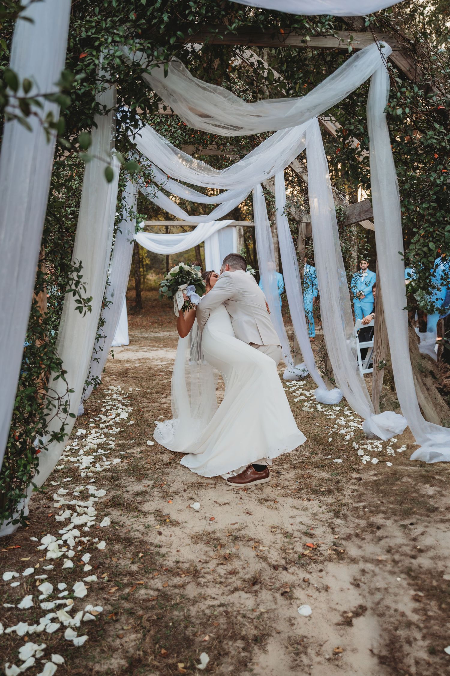 A couple in wedding attire kisses beneath elegant white fabric draping and scattered flower petals in Panama City Beach.