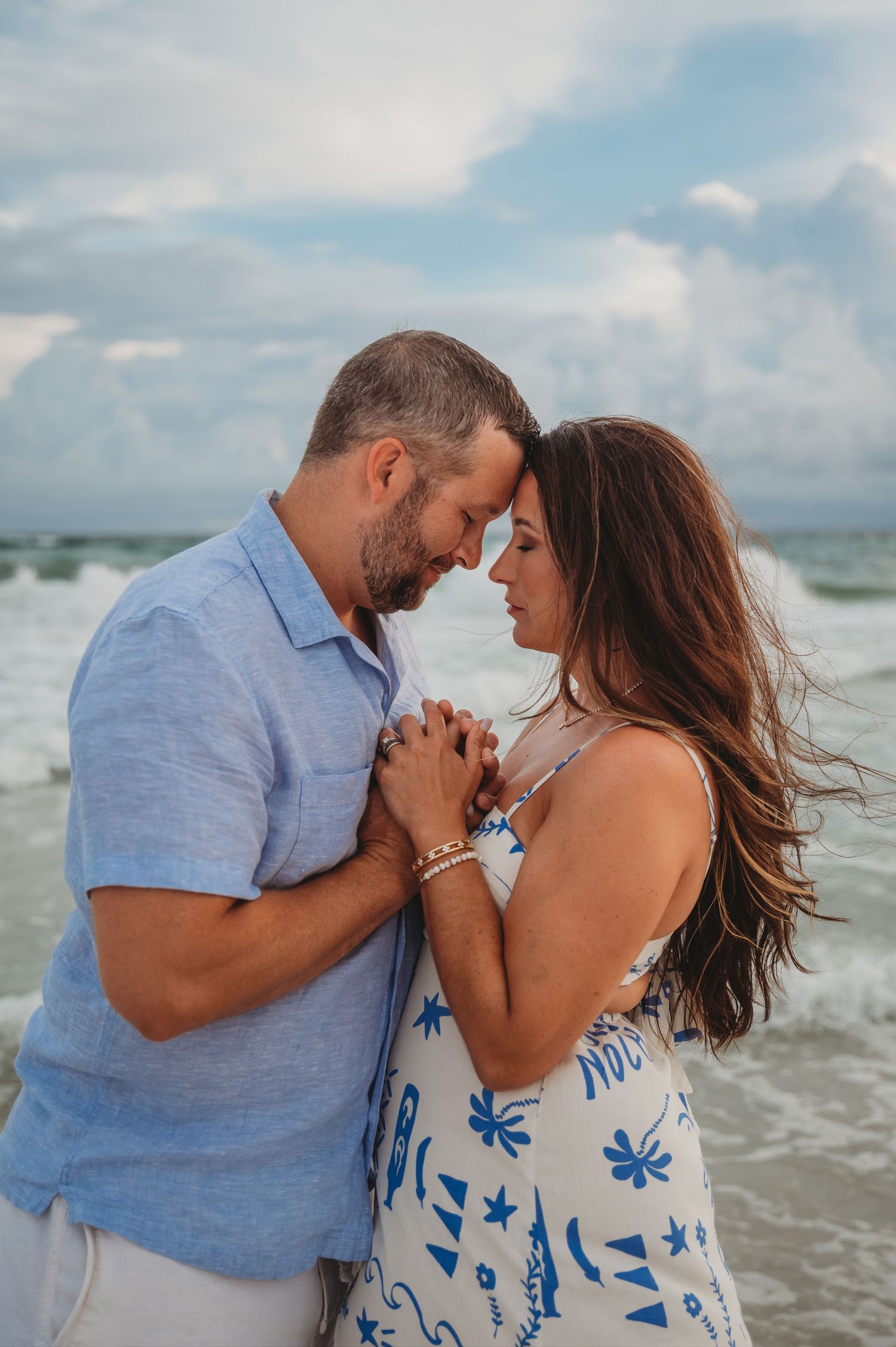 A romantic couple shares intimate moments on a beach with crashing waves and cloudy skies in the background in Panama City Beach.