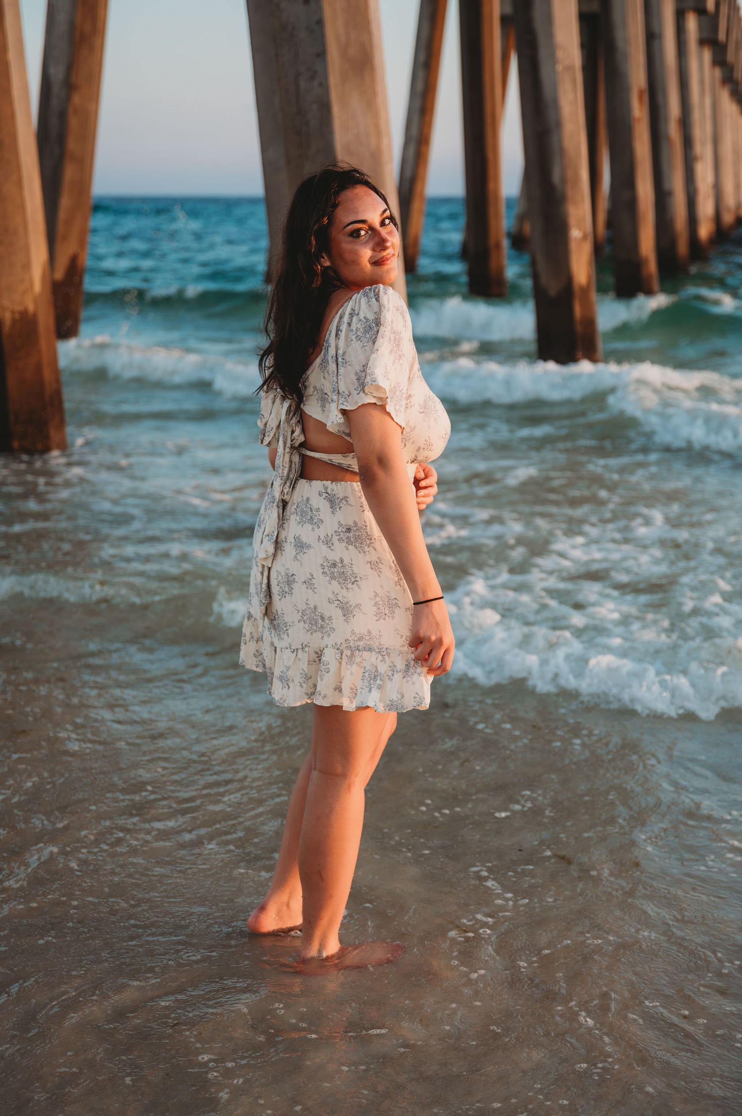 A white floral dress worn at sunset under a wooden pier as gentle waves lap at bare feet in turquoise waters in 30A.