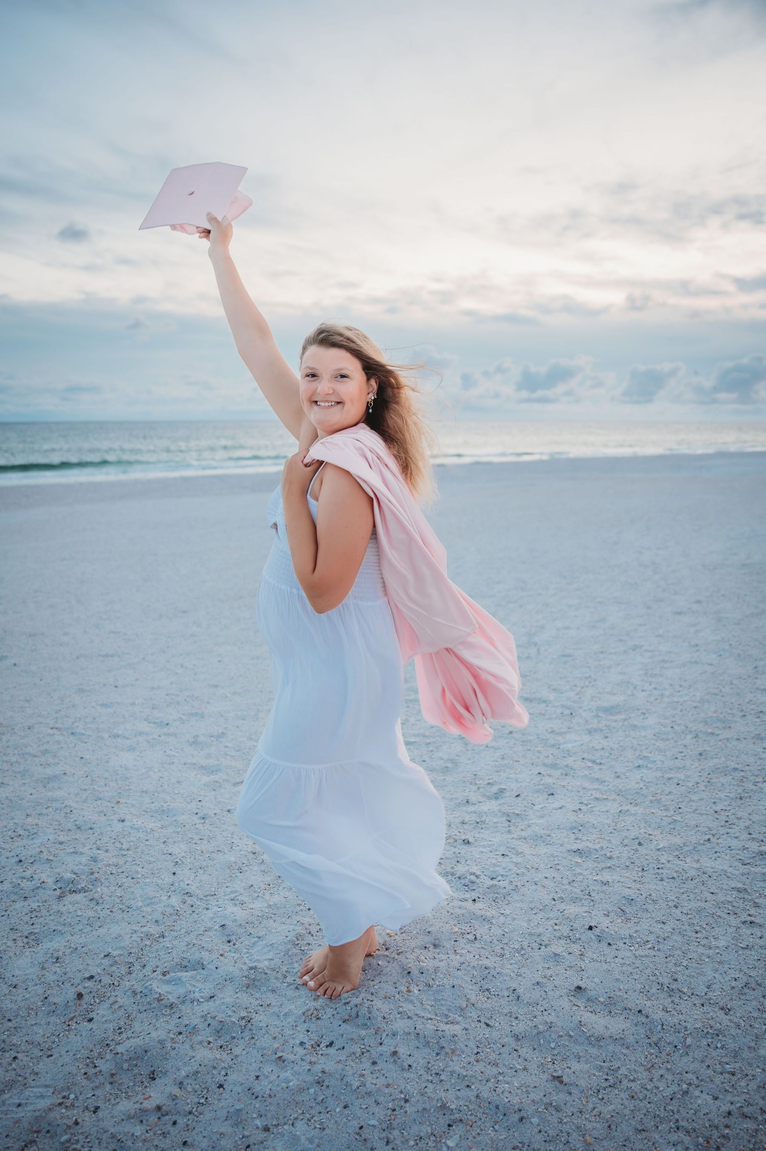 A senior in a flowing white dress dances joyfully on a beach at sunset while holding a pink graduation cap in Panama City Beach.