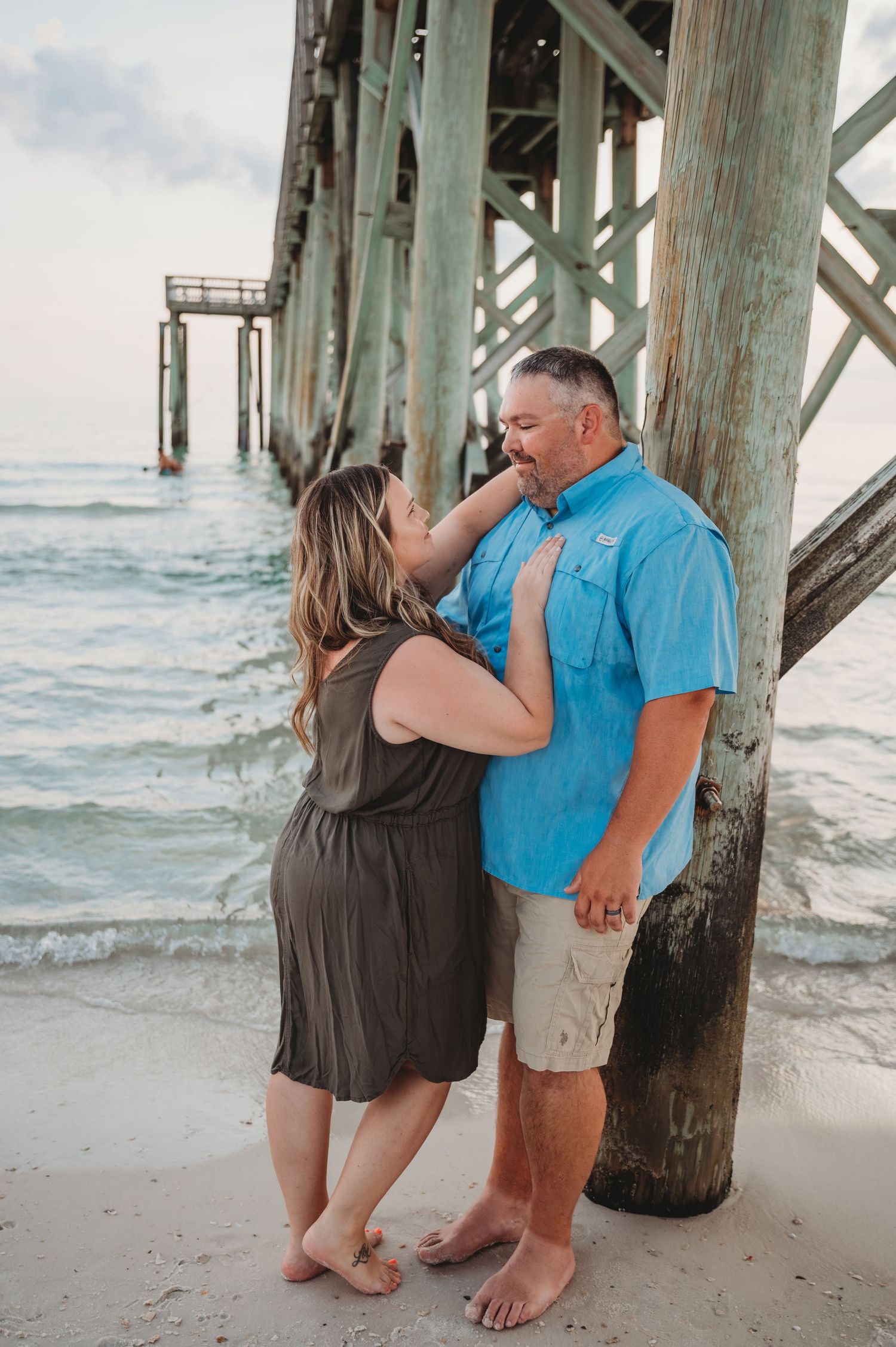 A romantic couple embraces under a wooden pier by the ocean during a beachy photo session in matching blue and brown outfits in Panama City.