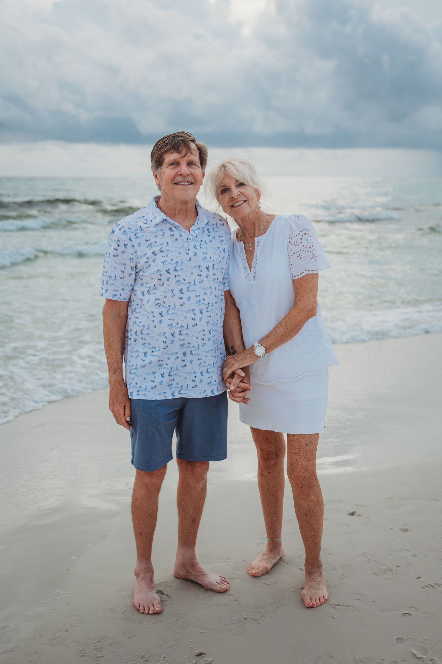 A loving couple stands barefoot on a beach at sunset holding hands with cloudy skies and ocean waves in the background in Panama City Beach.
