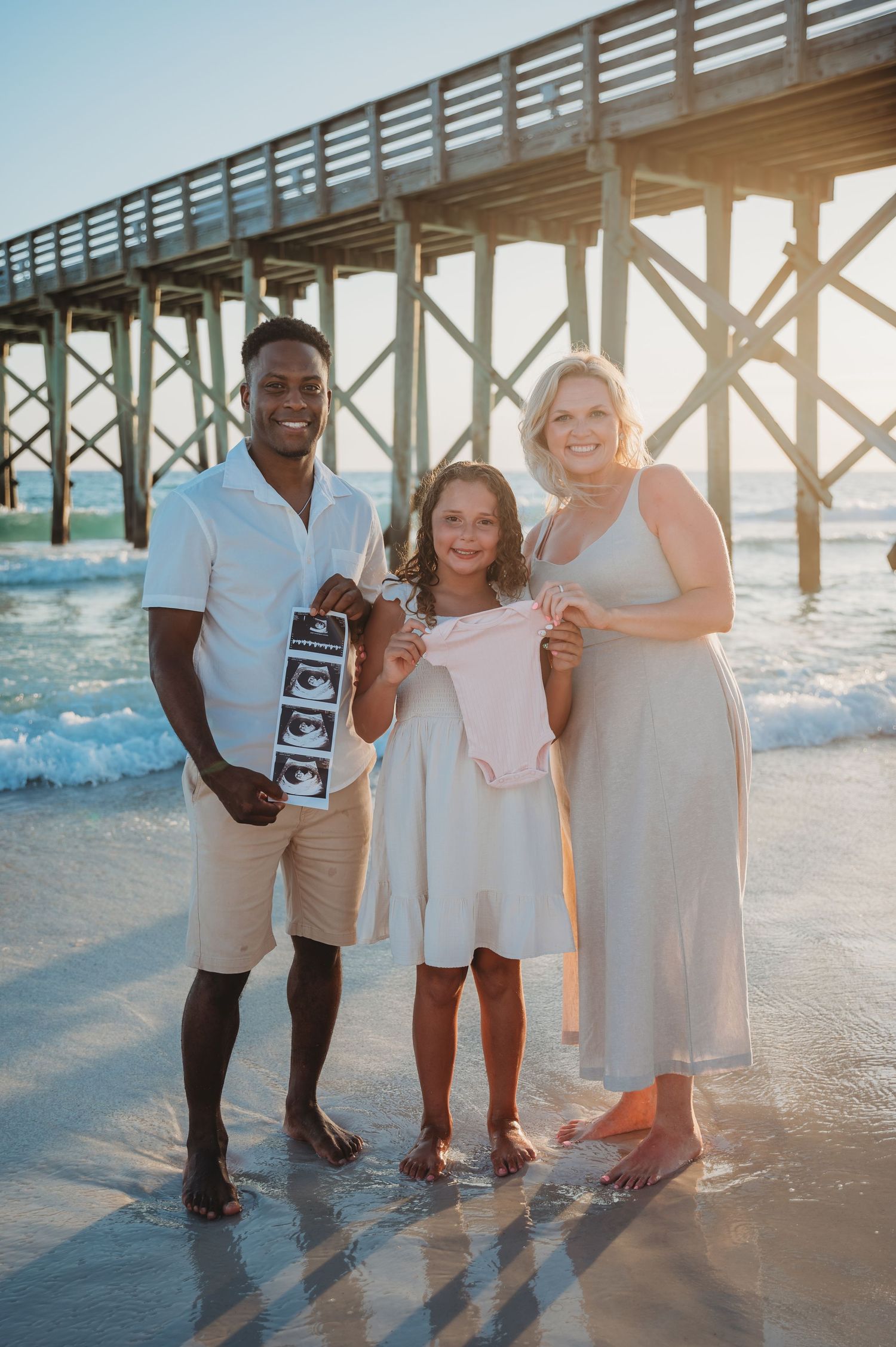 A family wearing white and beige outfits poses together on a beach near a wooden pier at sunset in Panama City Beach.