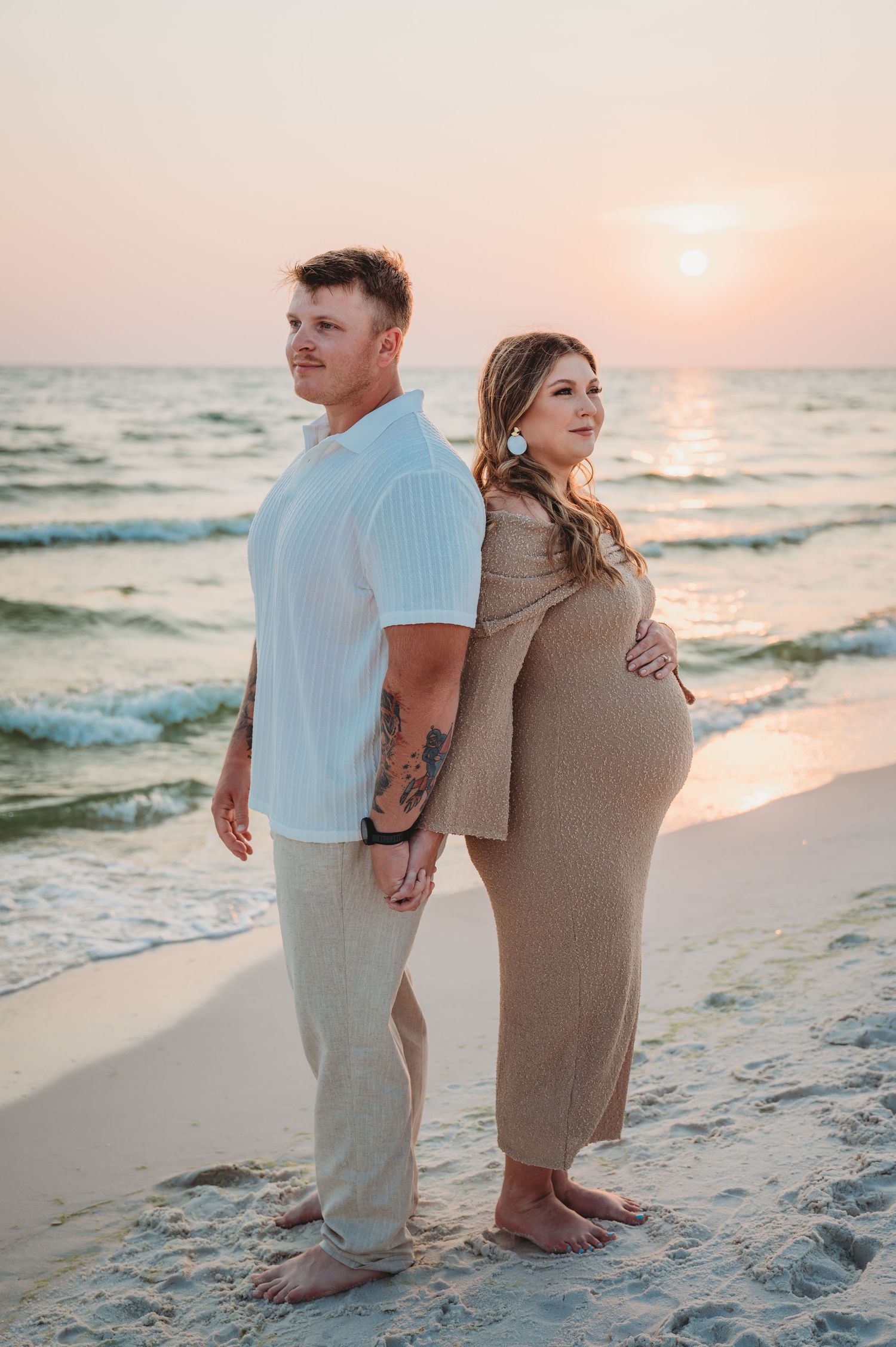 A couple stands together at sunset, watching the waves roll in along the shoreline during a maternity session in Laguna Beach.