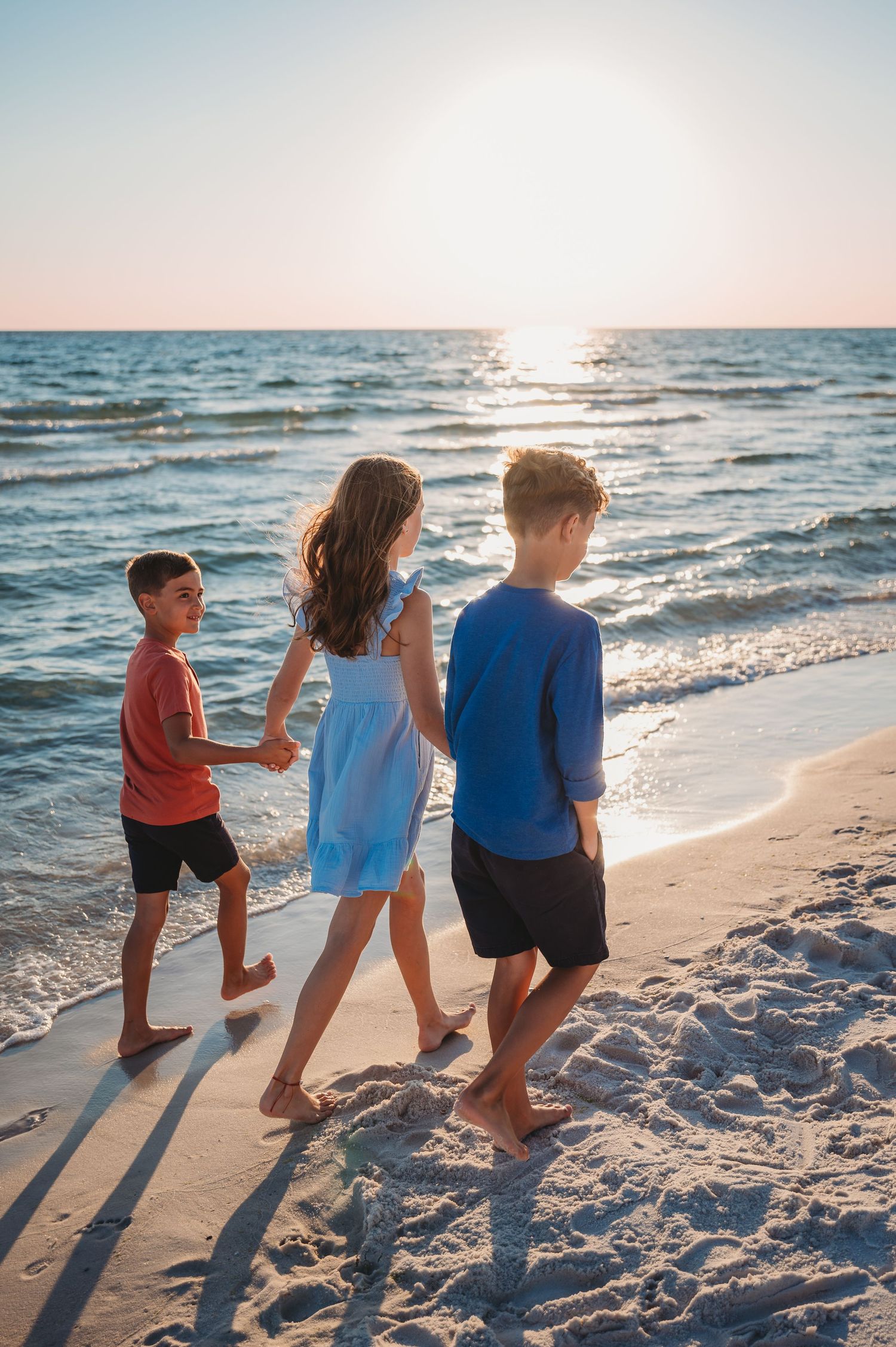Three children walking along a 30A beach at sunset with waves crashing on the shore and long shadows behind them.
