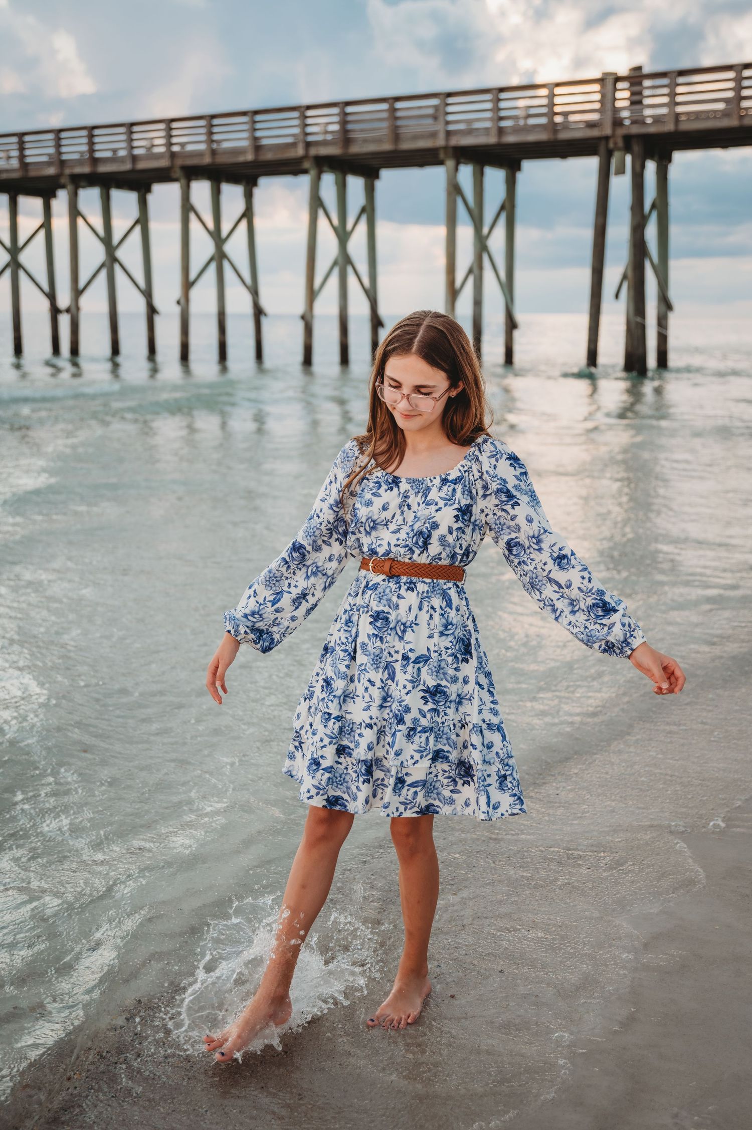Woman in blue and white floral dress playfully splashes in ocean waves near wooden pier at sunset in Panama City Beach.