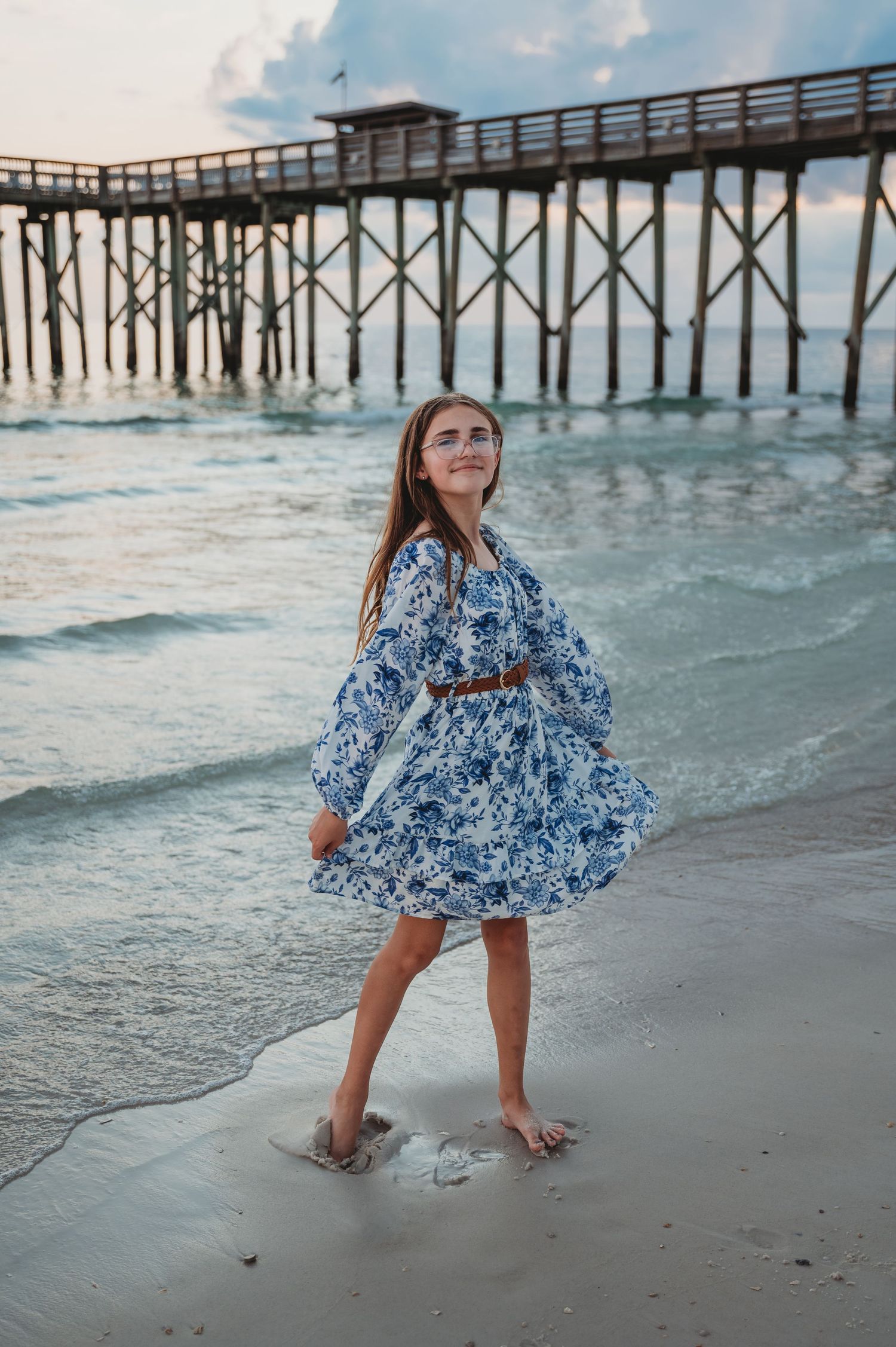 A young beachgoer in a flowy blue dress plays and twirls in the sand near a wooden pier at sunset in Panama City Beach.