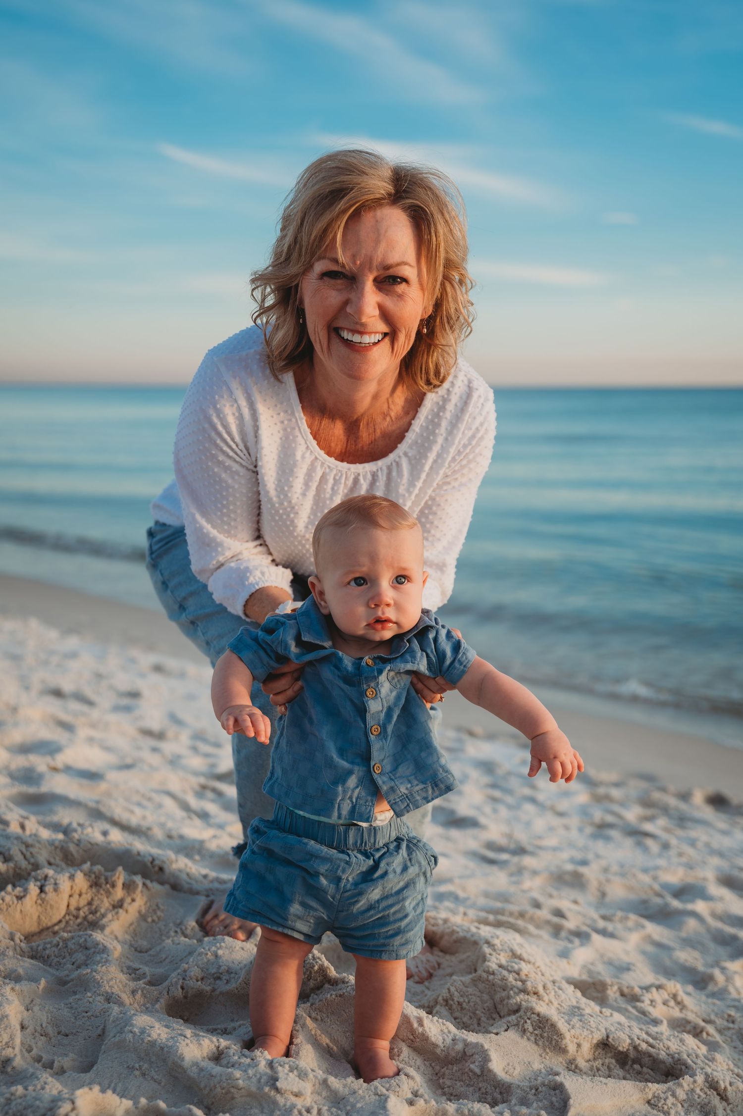 A toddler in denim overalls plays on a sunny white sand Panama City Beach while being supported by someone in a white sweater.