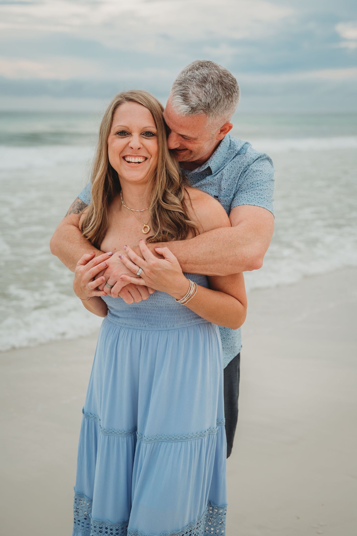 Couple in casual attire embraces on a cloudy beach with waves in the background in Panama City Beach.