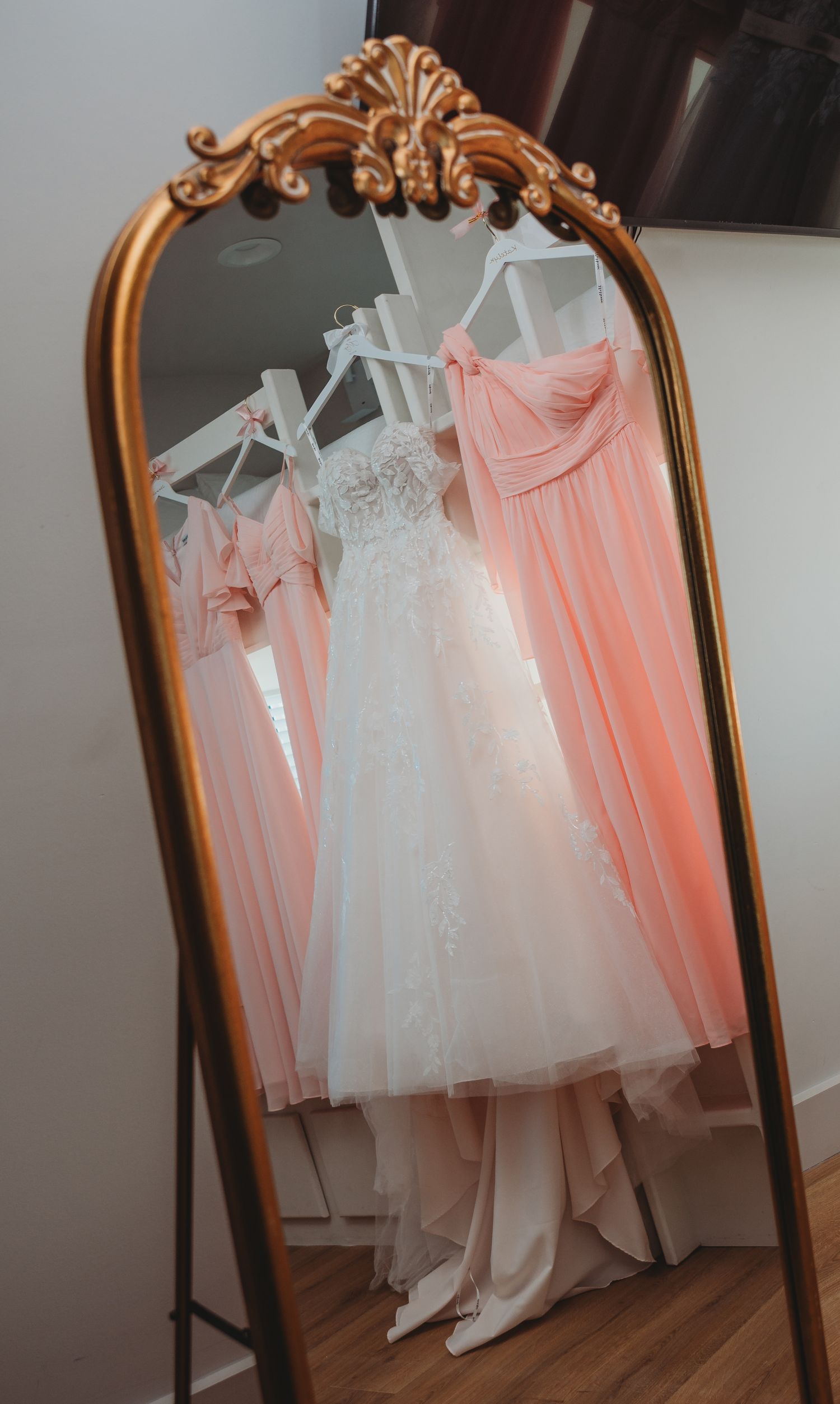 A collection of white and blush pink wedding dresses displayed on hangers reflected in an ornate gold-framed mirror during a 30A wedding.