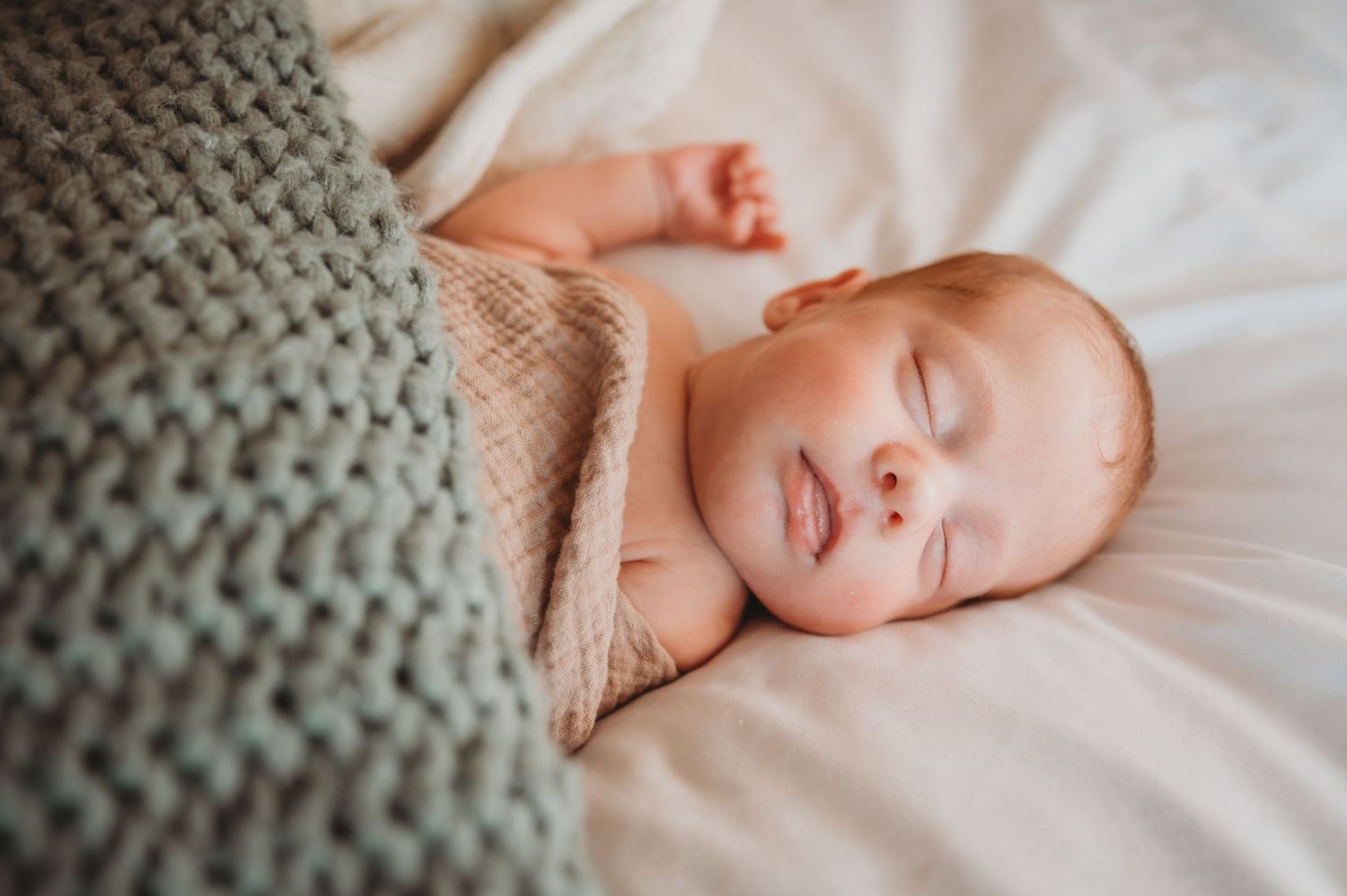 A newborn baby rests peacefully on soft white bedding with a knitted gray blanket visible during a newborn session in Panama City.