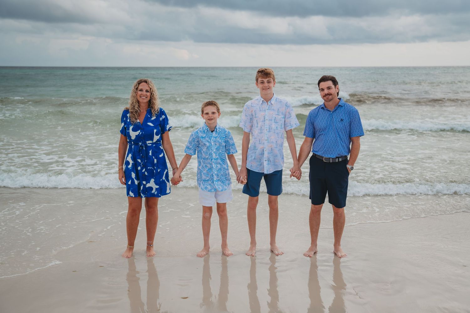 A group of people in coordinating blue beachwear smile while standing together on a sandy beach at the ocean's edge in Panama City Beach.