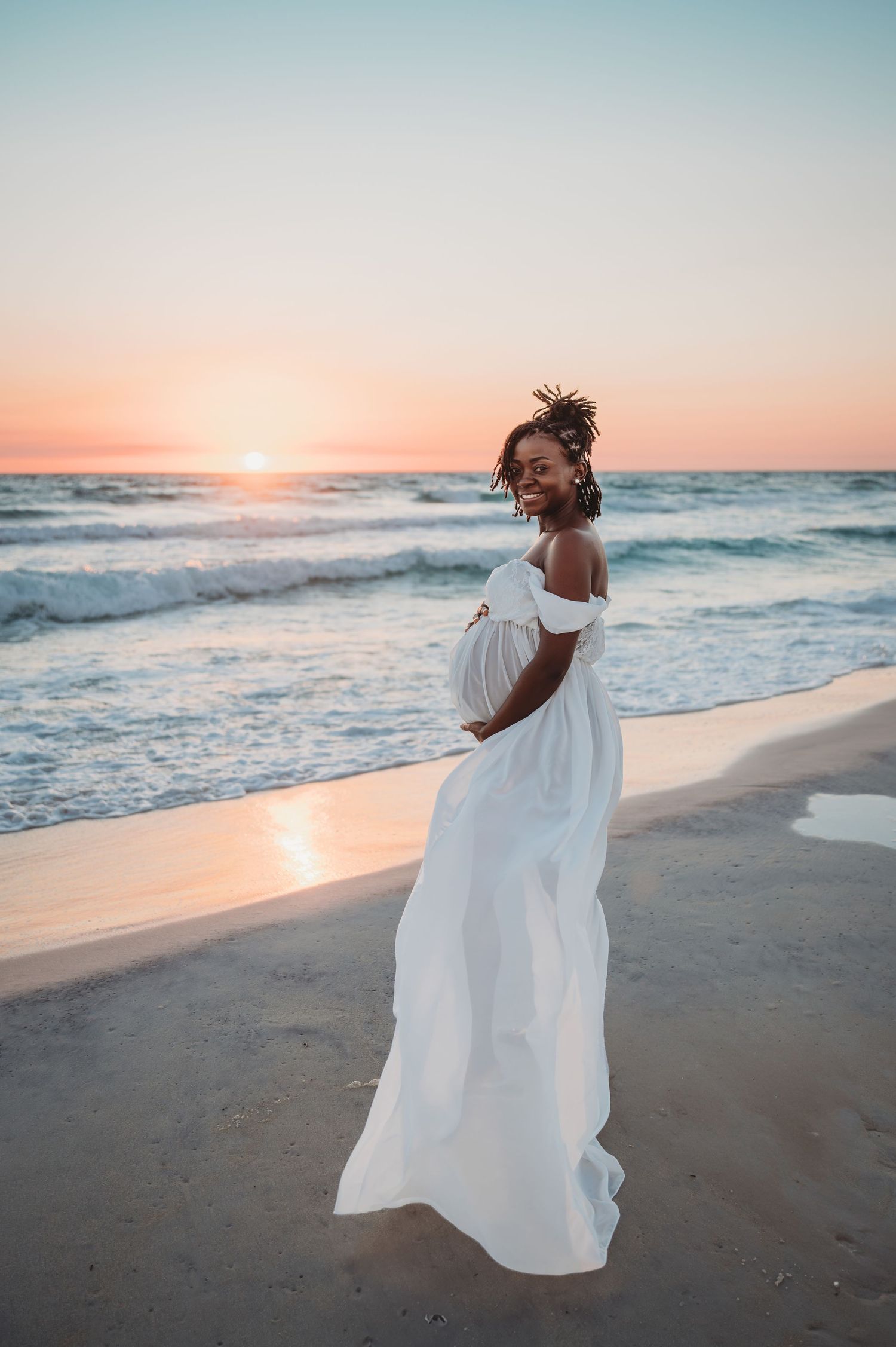 A pregnant woman in a flowing white dress stands on a 30A beach at sunset as waves crash behind her.