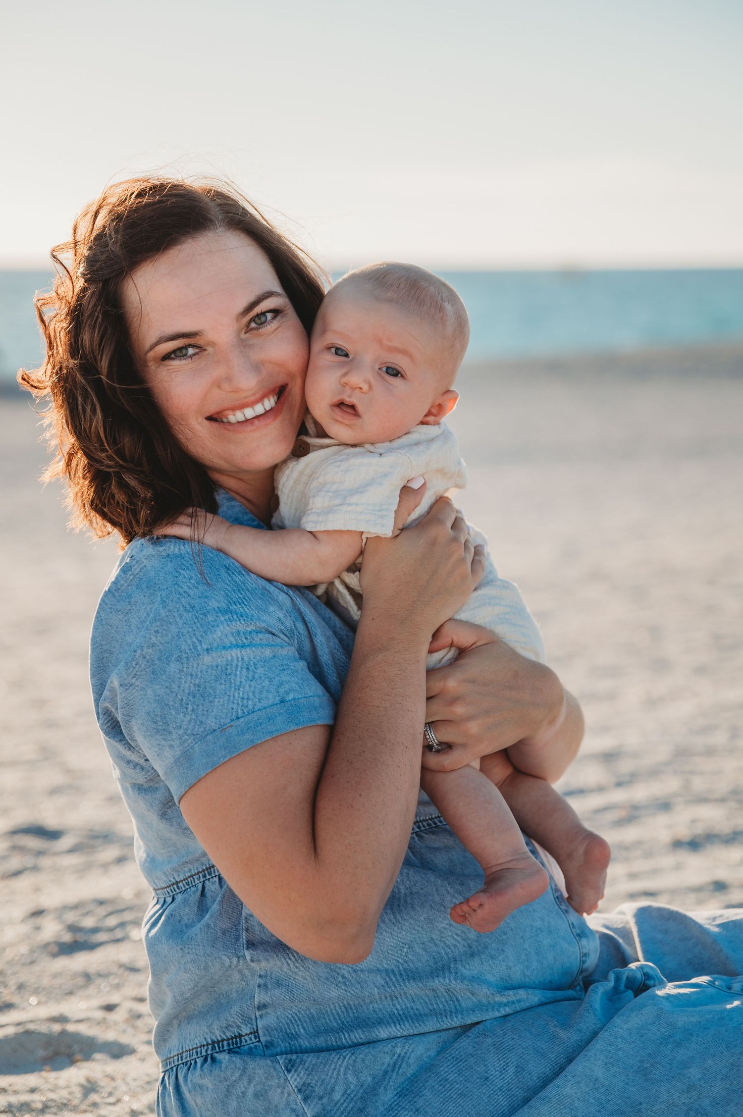 A woman in a blue shirt holds and cuddles with a baby on a sunny 30A beach during a tender moment.