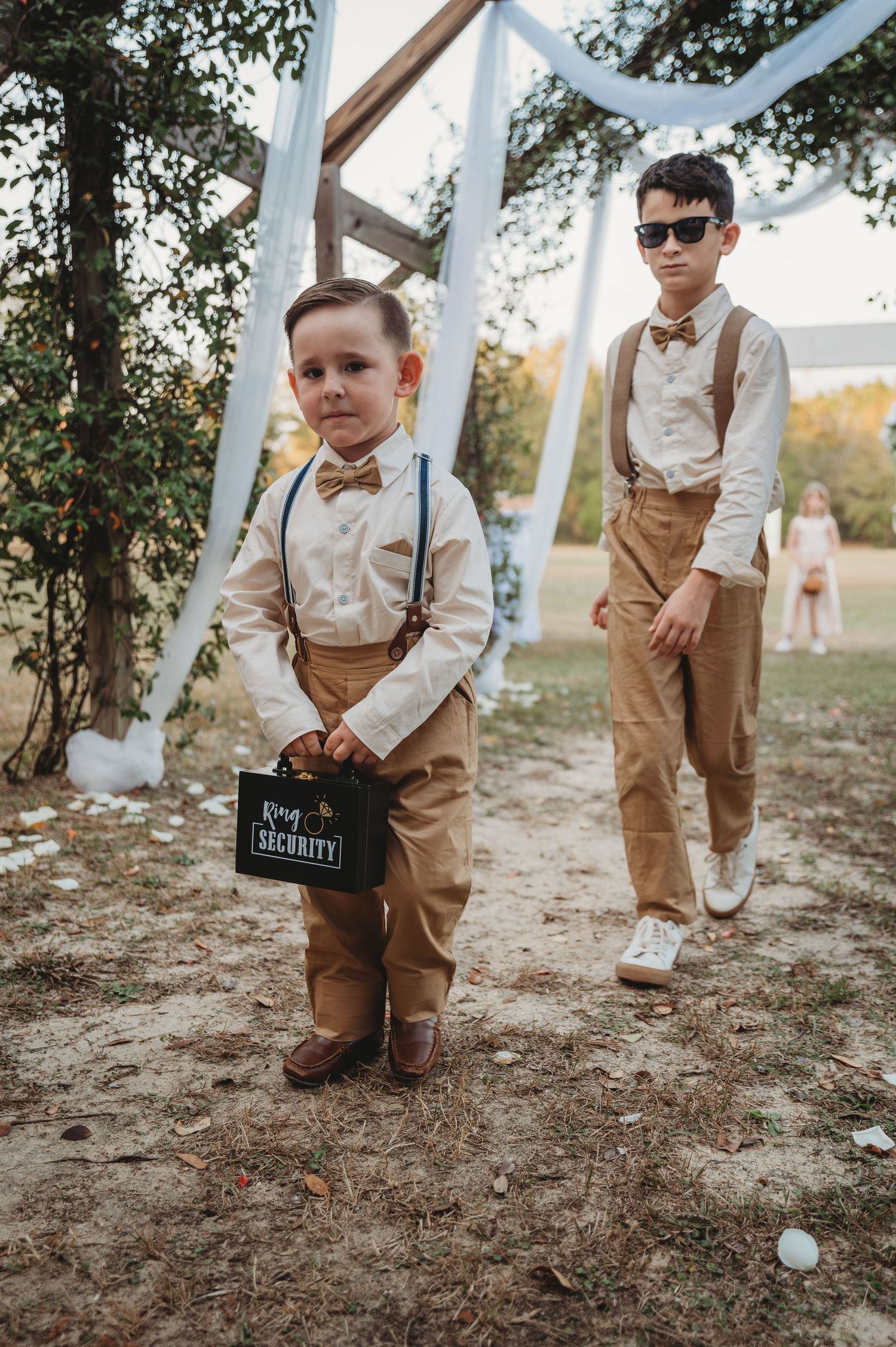 Two ring bearers in matching tan suspenders and bow ties stand under white drapery at an outdoor wedding ceremony in Panama City Beach.
