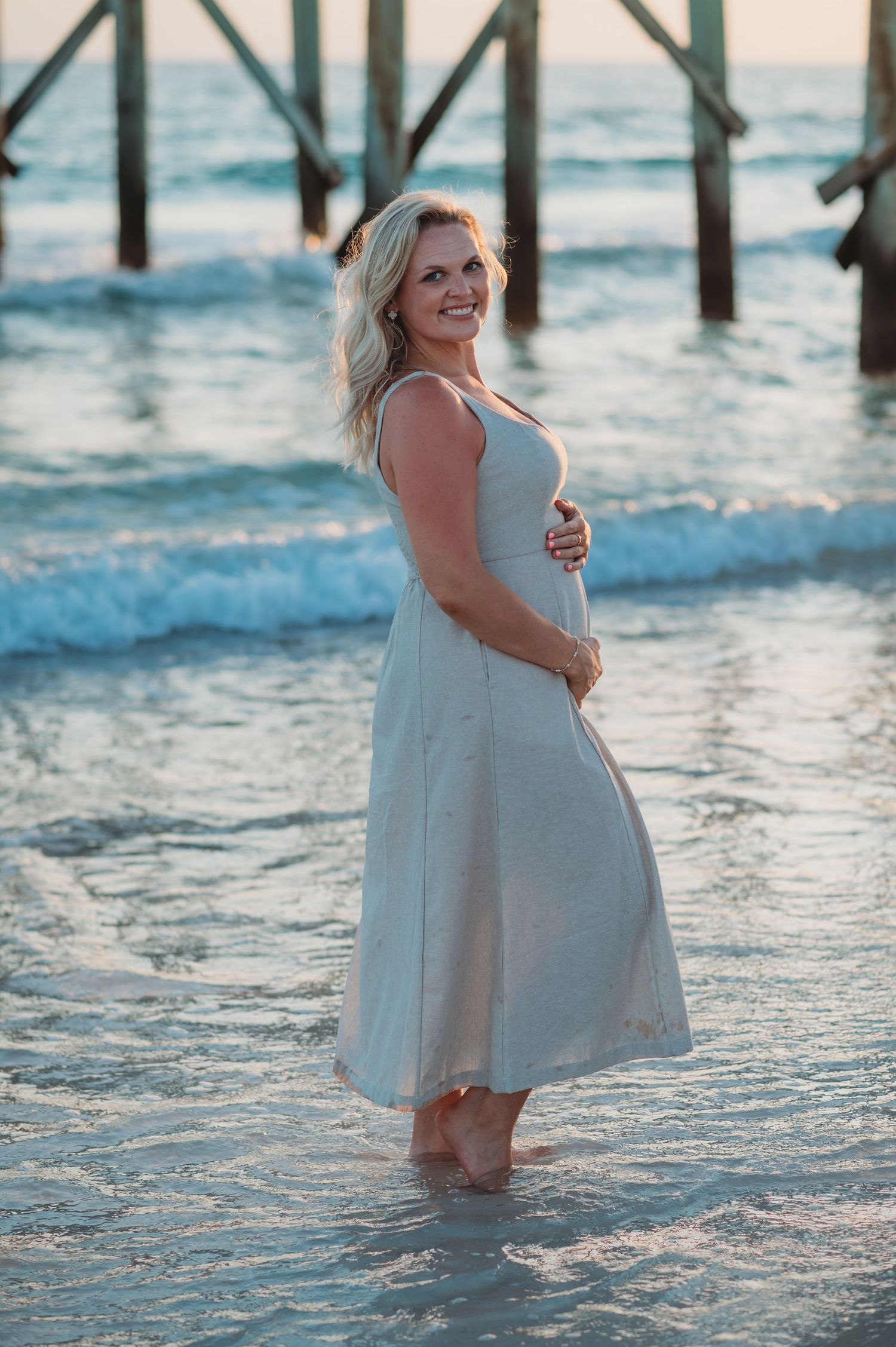 A beachgoer in a white dress poses playfully near waves under a wooden pier in Panama City Beach.