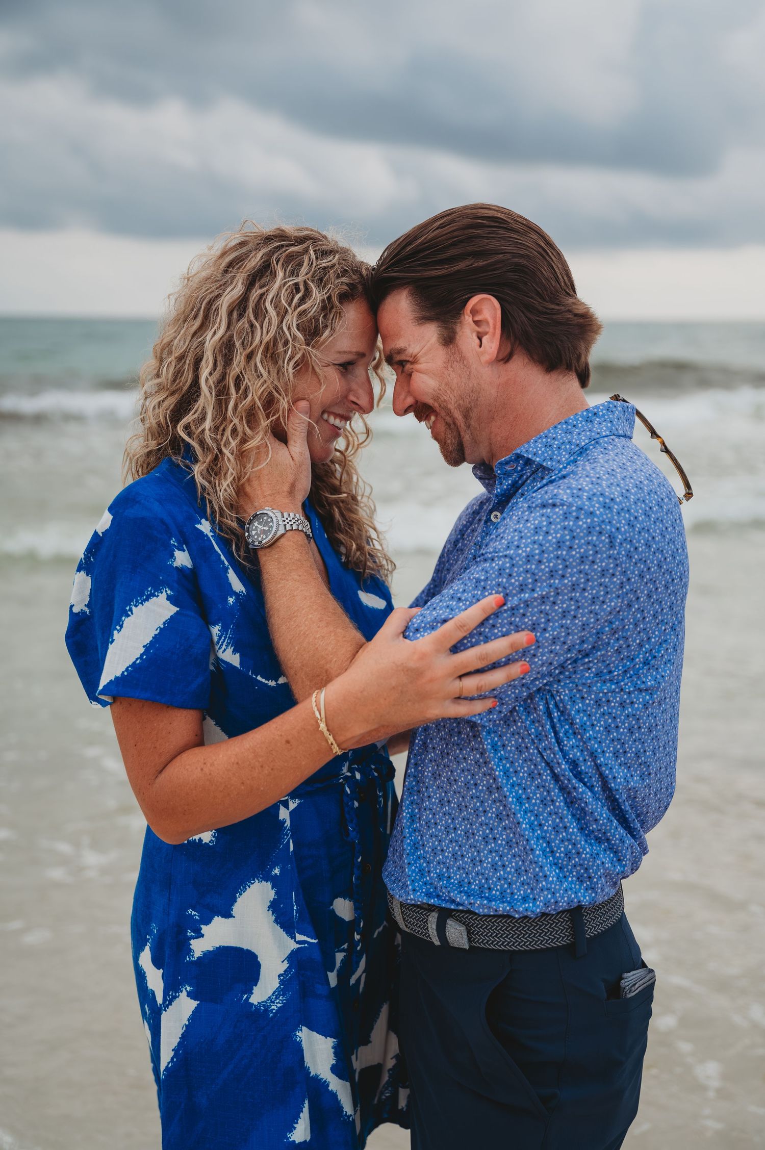 A couple in blue attire share intimate moments on a beach with ocean waves and cloudy skies in the background in Panama City Beach.