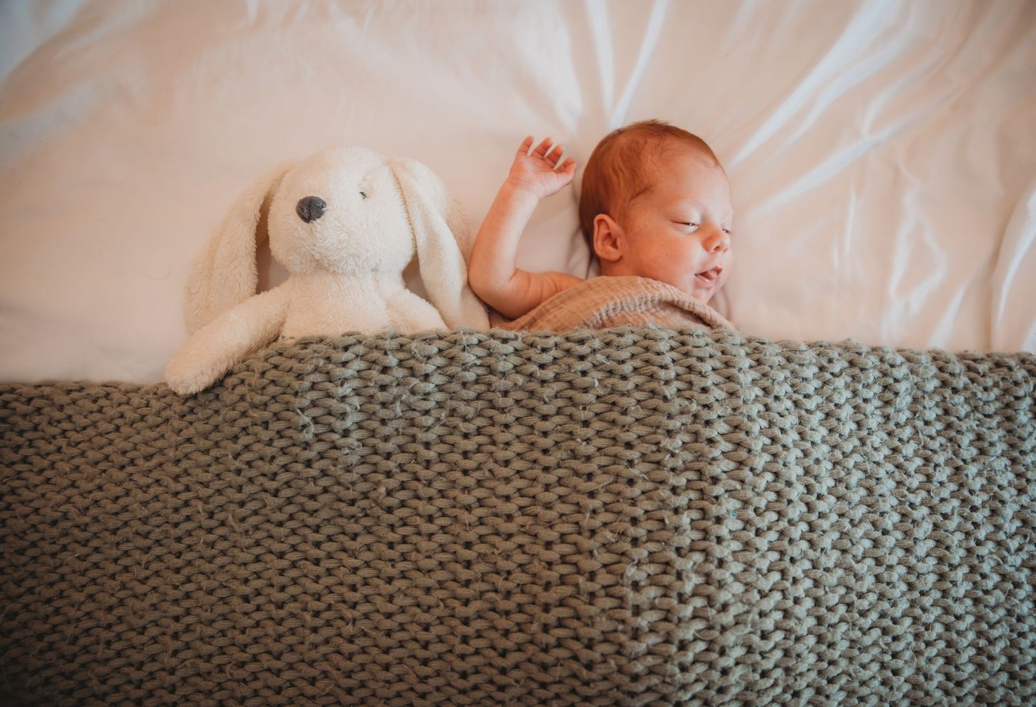 Newborn baby resting on white bedding with knit gray blanket and plush toy companion during a newborn session in Panama City.