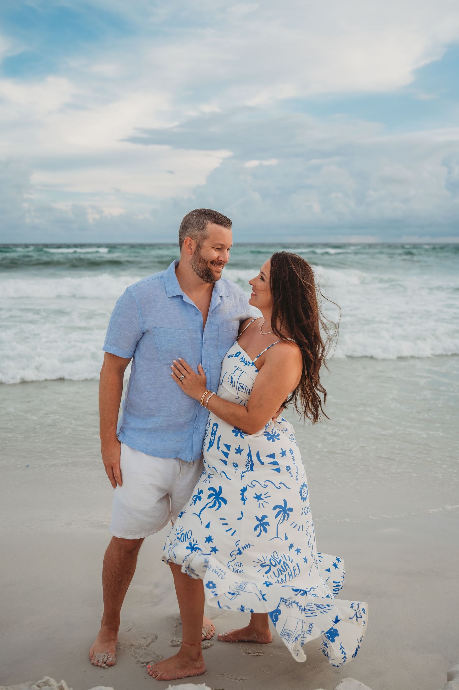 A couple in coordinating blue and white outfits share romantic moments while walking along a sunny beach shoreline in Panama City Beach.