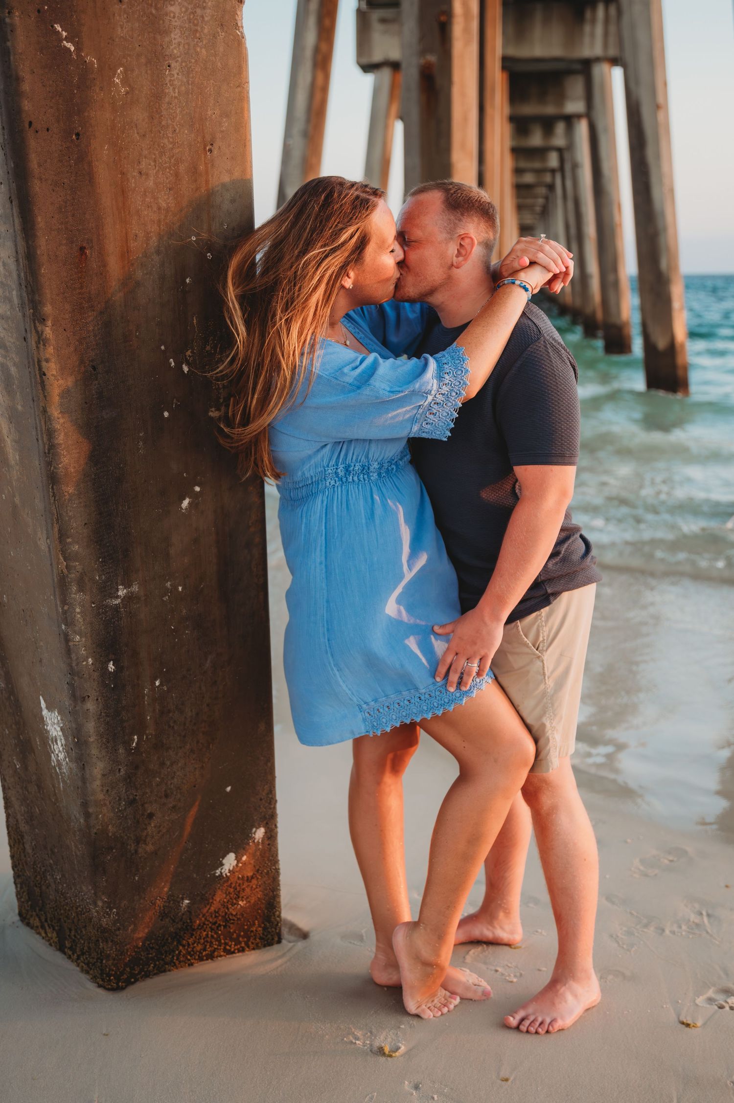A couple shares a romantic moment beneath a pier on the beach at sunset, with ocean waves visible in the background in Panama City Beach.