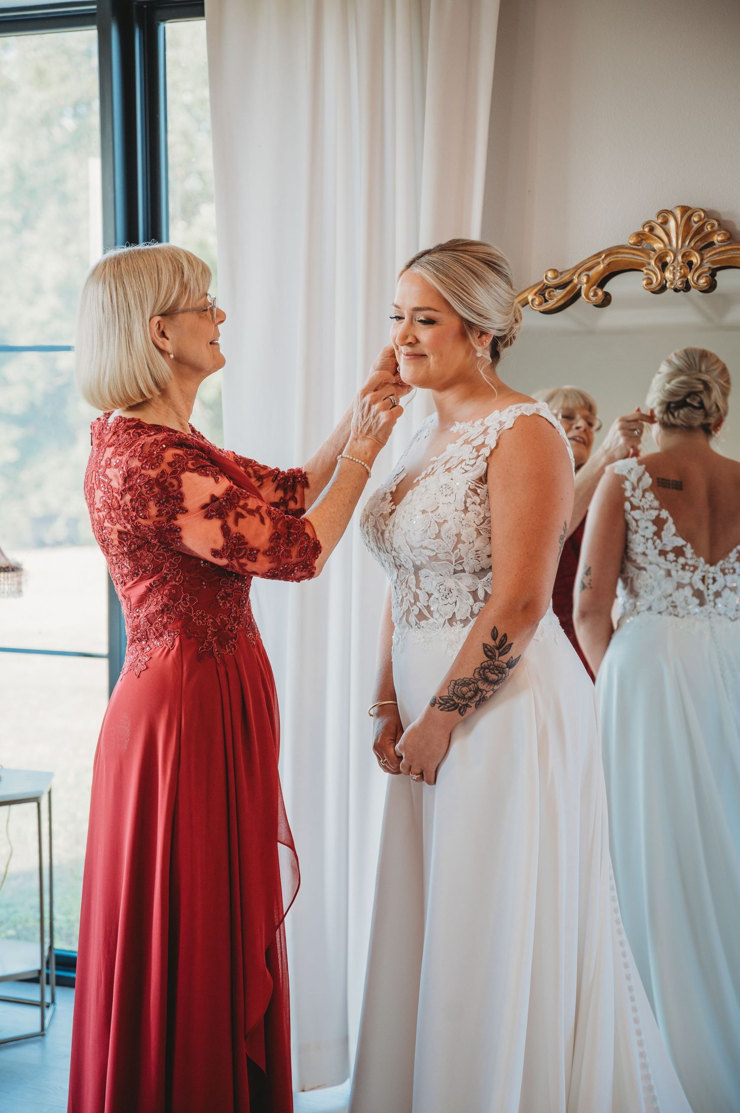 A woman in a red gown helps adjust makeup on someone wearing a white lace wedding dress in a sunlit room from a Panama City Beach wedding.