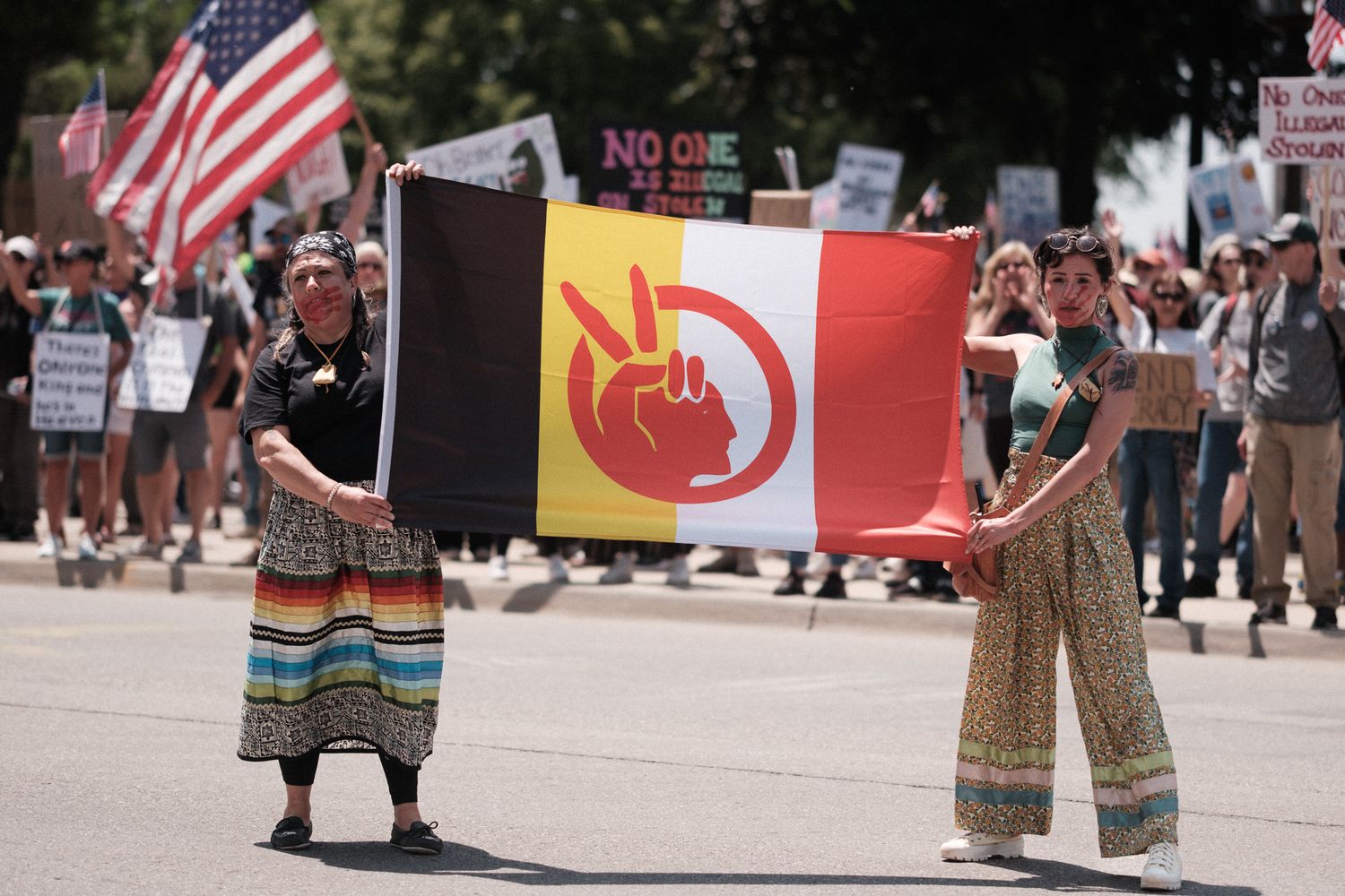 Protesters hold a red and black Indigenous rights flag at a demonstration with American flags in the background.