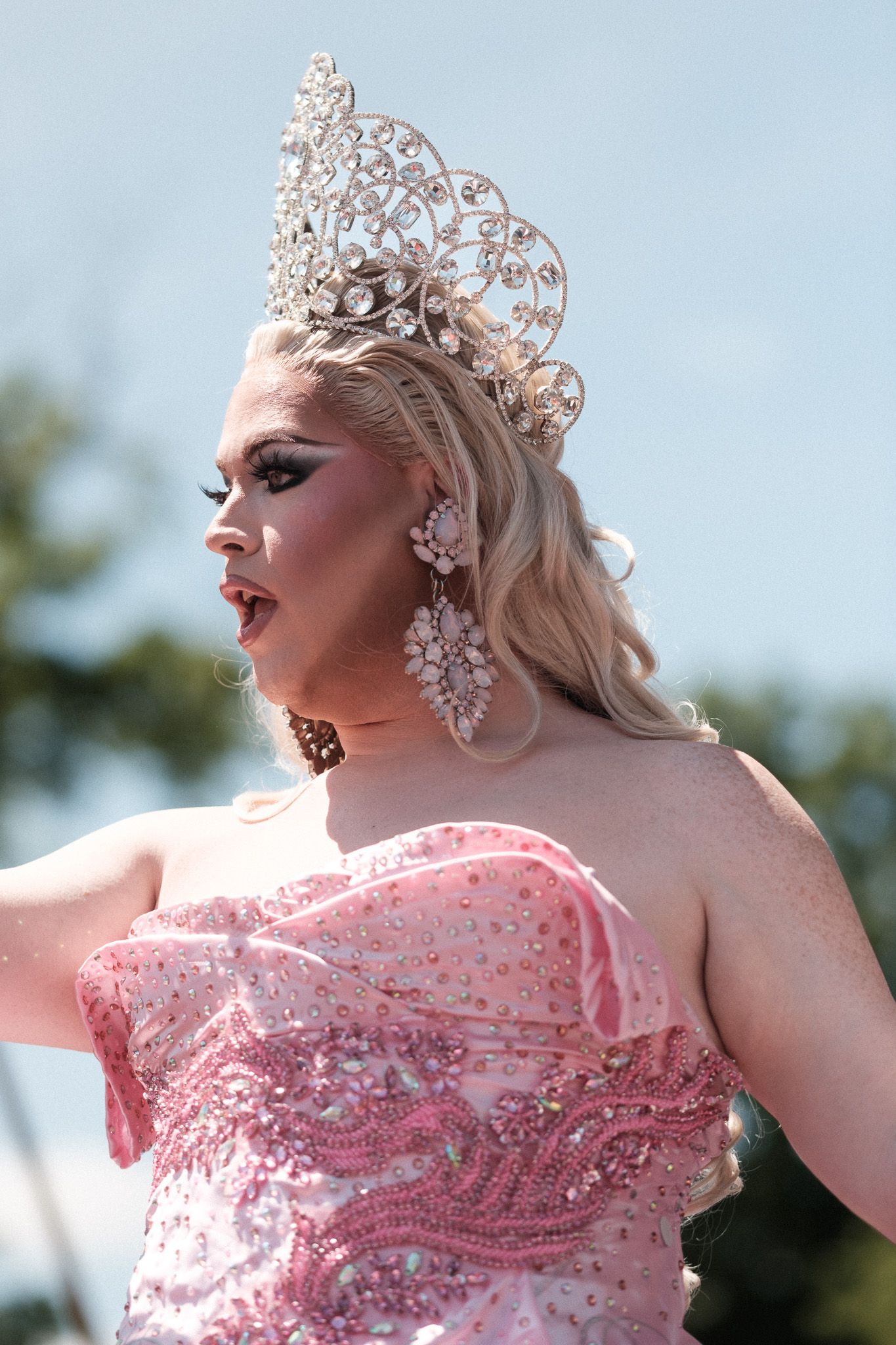 A performer in a glittering pink gown and ornate crown poses regally outdoors against a bright blue sky background.