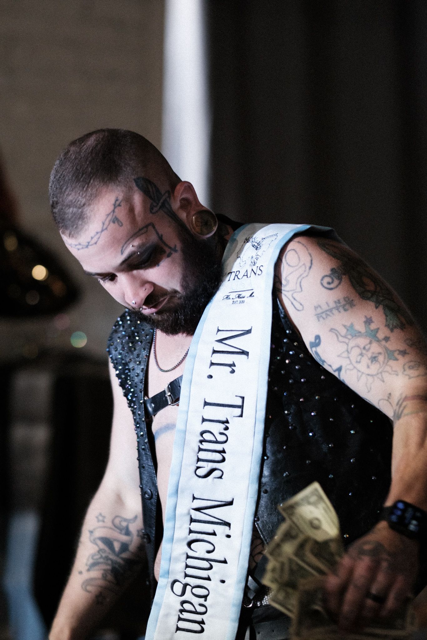 Tattooed performer wearing 'Mr Trans Michigan' sash smiles during competition.