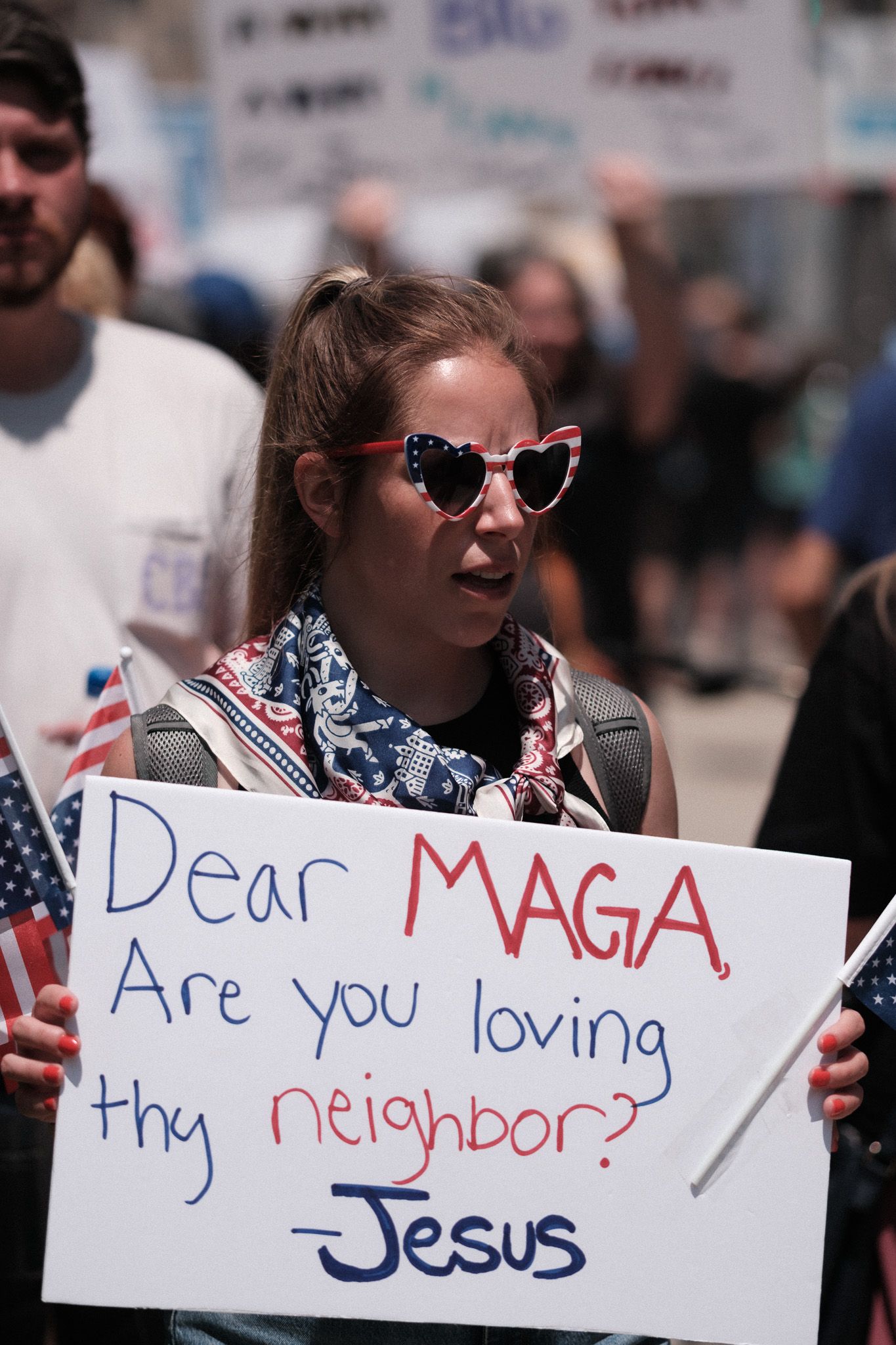 A young protester wearing heart sunglasses holds a sign questioning MAGA supporters about loving thy neighbors.