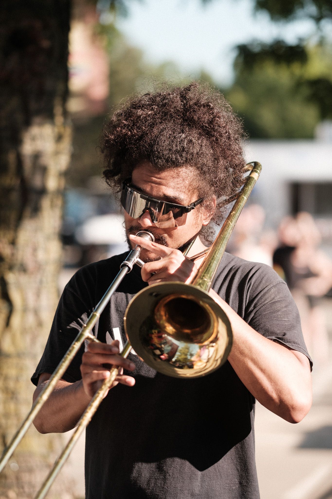 Musician with curly hair plays trombone outdoors while wearing sunglasses and a black t-shirt.
