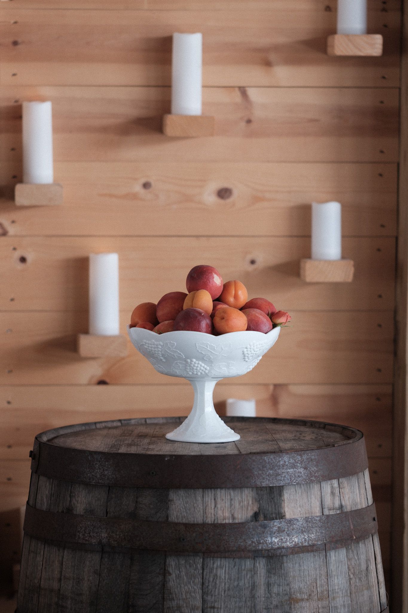 Peaches displayed in a white pedestal bowl on top of a rustic wooden barrel with wooden wall paneling in the background.