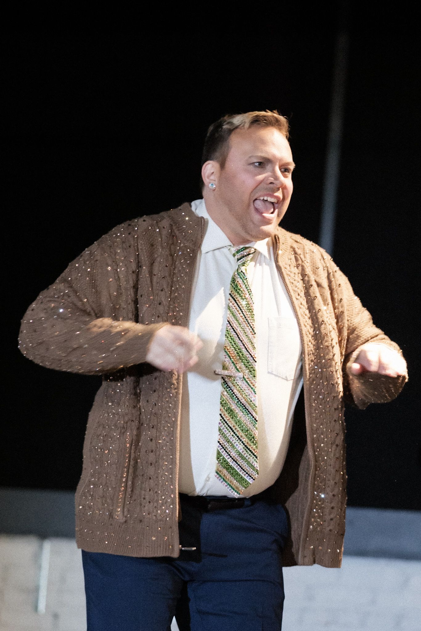 Energetic performer in brown cardigan and striped tie gesturing dramatically.