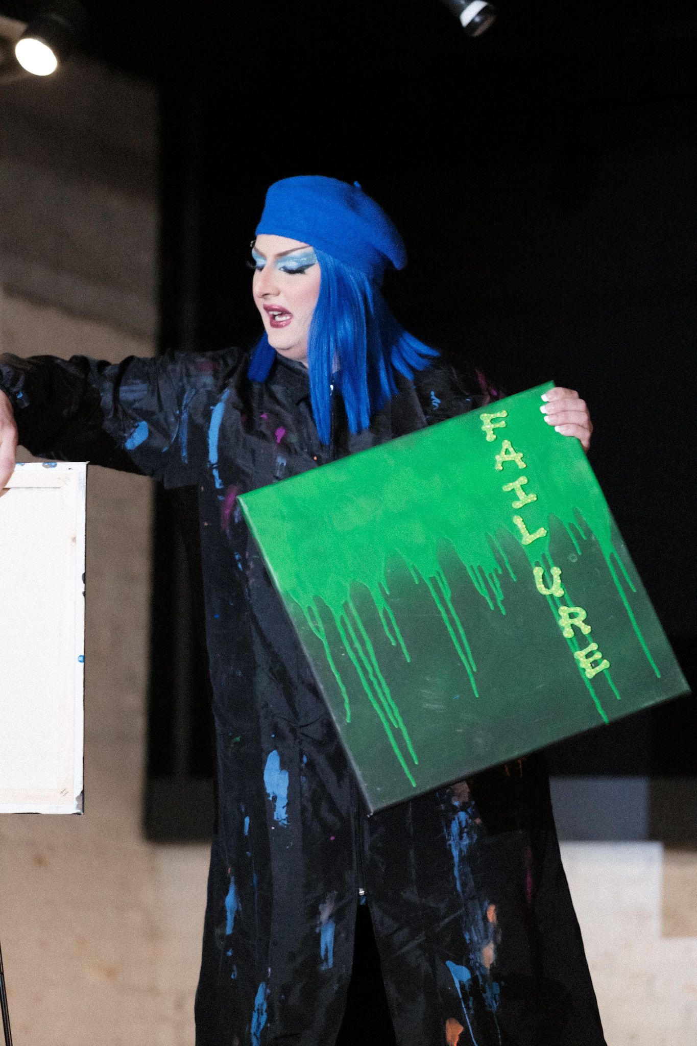 Performer in blue hair and dark outfit holding a bright green dripping artwork sign.