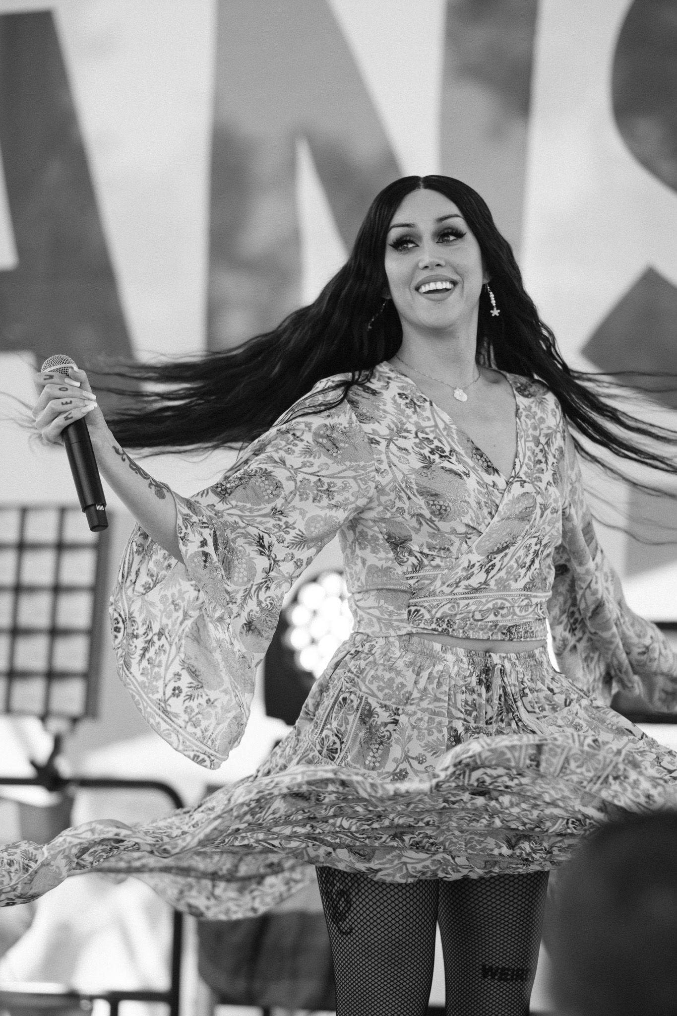 A performer in a flowing silver lace dress with bell sleeves dances joyfully on stage with long dark hair flowing.
