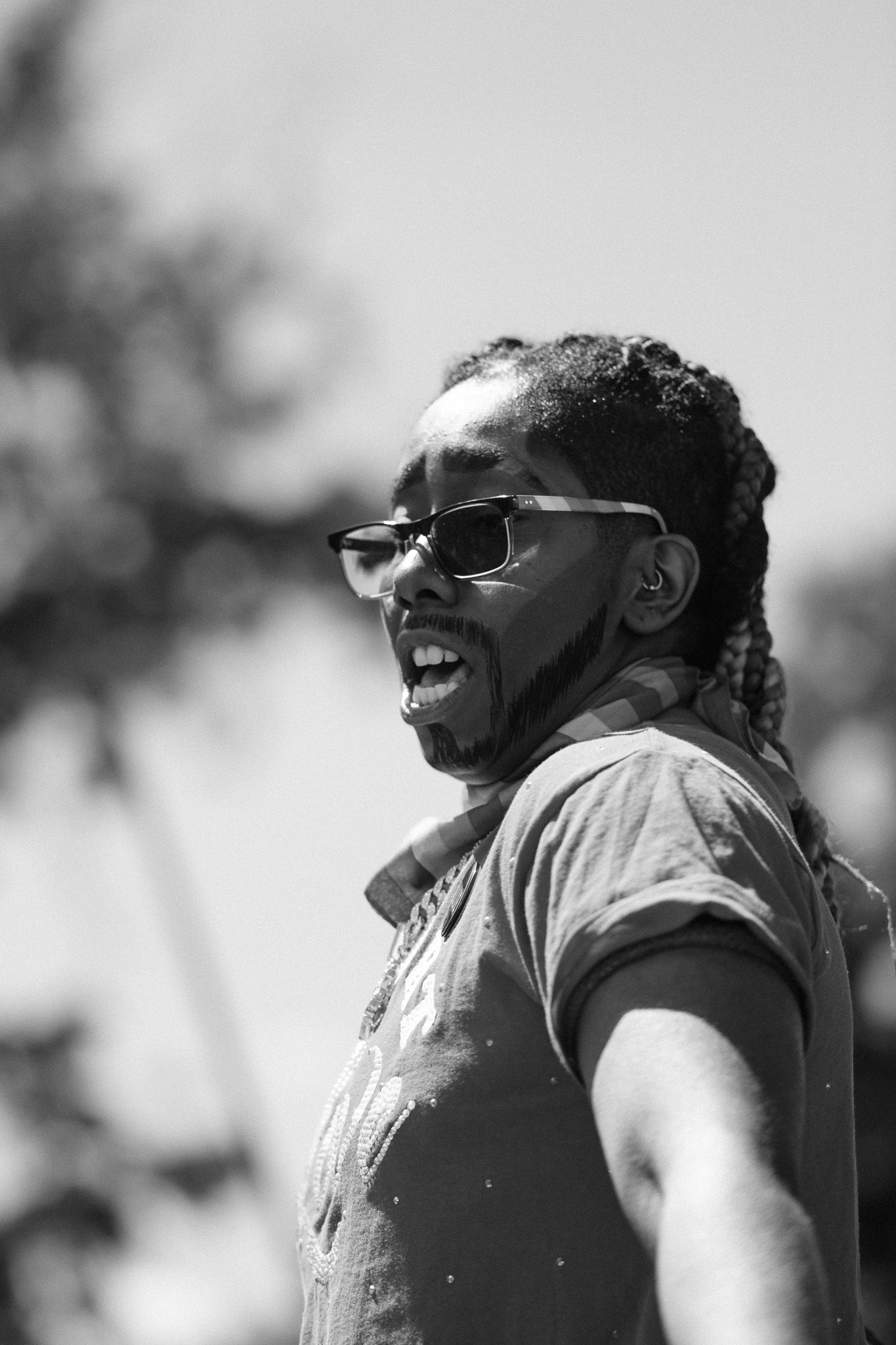 Black and white portrait of a smiling person wearing glasses at an outdoor event.